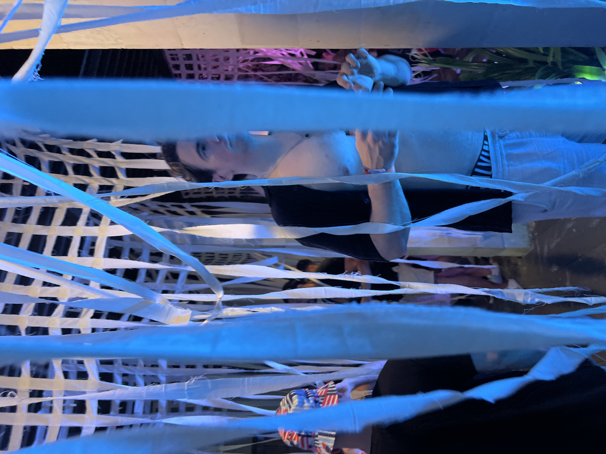 The image, oriented vertically, displays a person suspended within a dense, crisscrossing web of white, ribbon-like straps or fabric. This individual has dark hair and light skin, wearing a dark top (possibly a sports bra or swimsuit) and light-colored shorts or trousers. Their eyes are open, looking towards the top of the frame. A thin necklace adorns their neck, and a wristband is visible on their left wrist.
Behind the prominent white straps, a black grid-like structure is discernible, partially obscured by the straps themselves. The scene is illuminated by dramatic blue light, which casts a glow on many of the straps, making them appear luminous blue. In the background, there are glimpses of pink and purple lighting, along with some green foliage and a dark, possibly woven basket.
A second person is partially visible at the bottom of the original image, holding an item patterned with red, white, and blue stripes and stars, reminiscent of the American flag. The setting appears to be an indoor or sheltered environment, likely at night or under artificial conditions, given the moody, colored lighting. It suggests an artistic installation, a themed event, or a performance space. No visible text is present in the image.