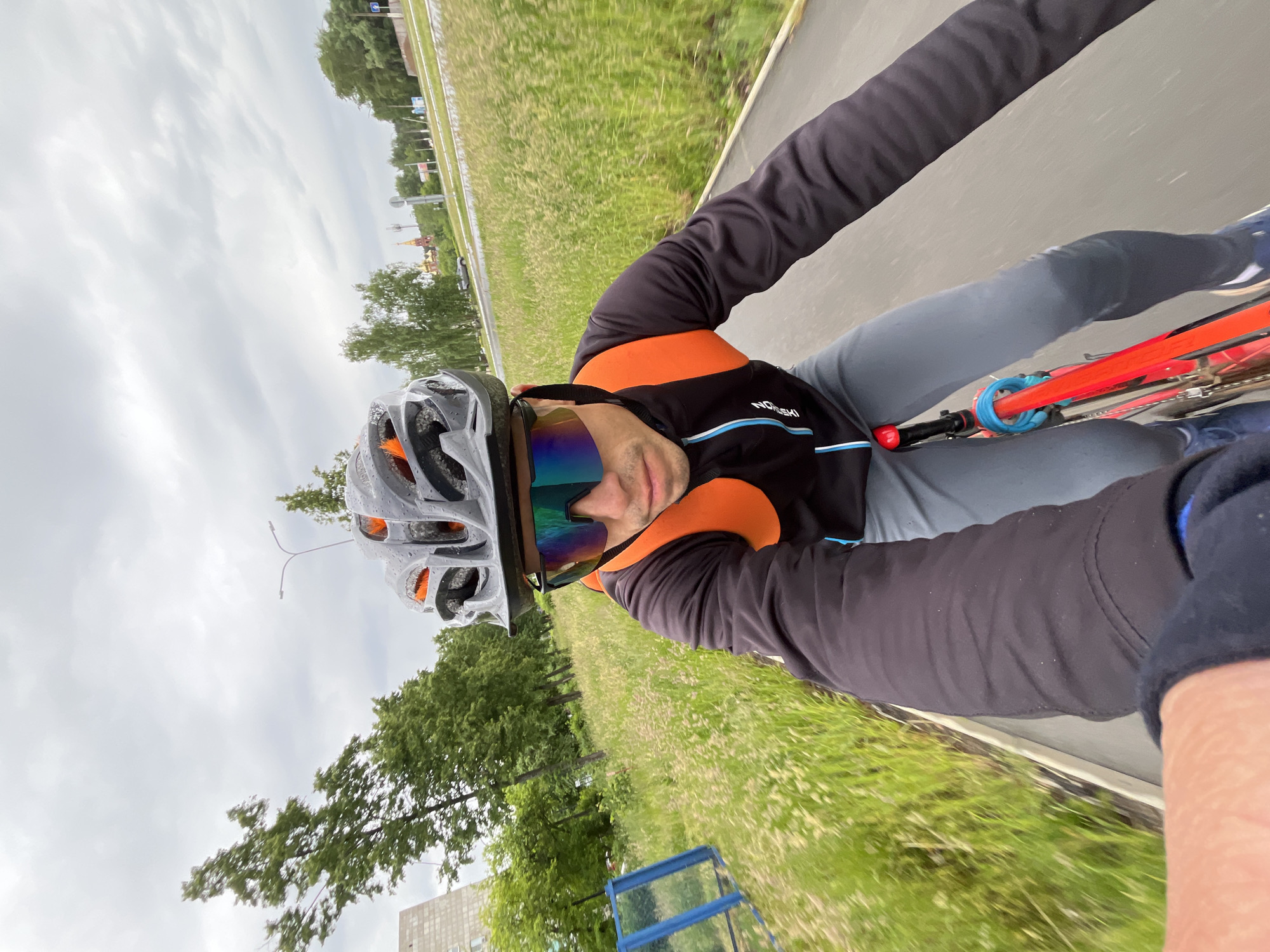 The image is a rotated, first-person perspective (selfie) capturing a person cycling outdoors on an asphalt path. The cyclist wears a grey helmet with visible water droplets, mirrored sunglasses reflecting blues and oranges, and a black long-sleeved cycling top featuring orange shoulders, a blue stripe, and the text "NOESKI" on the chest. Grey cycling pants are also visible, along with a red bicycle frame that has a blue lock or cable wrapped around its top tube.
The path is flanked by wide grassy verges; the one on the right shows motion blur, indicating movement. Beyond the grass, there are trees and distant urban structures, including buildings and a street light pole. A faint, distinctive red-roofed building with a spire can be seen in the far distance on the right, and a blue metal structure (possibly a fence or bench) is in the mid-ground on the left. The sky is entirely overcast and cloudy, suggesting a dull day, possibly after recent rain given the moisture on the helmet. The scene is set in Chaikovsky, Russia.