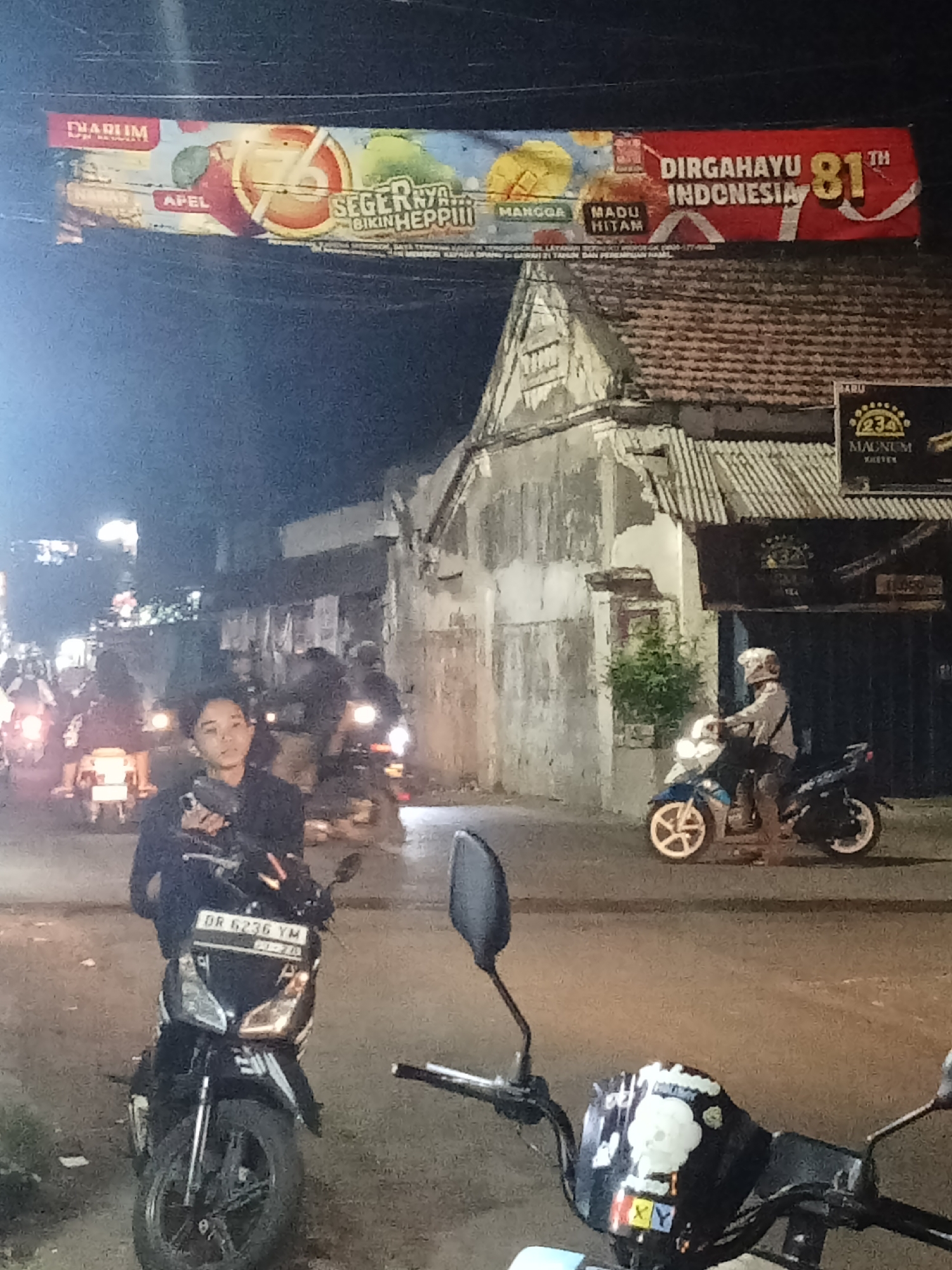 The image depicts a nighttime street scene in Peterongan, Indonesia, likely a commercial area given the banner and signage. A large banner stretches across the top of the frame, advertising "Dihatuyu Indonesia 81st". Below the banner, a row of buildings line the street, some with tiled roofs and weathered facades. Several motorcycles are parked or in motion. In the foreground, a woman wearing dark clothing stands next to a black motorcycle with the license plate "DR 6236 YM". To the right, another individual on a blue and white motorcycle is visible. Other motorcycles and people are present in the background, illuminated by streetlights and the headlights of vehicles. The overall atmosphere is that of a busy, possibly bustling, urban environment at night.