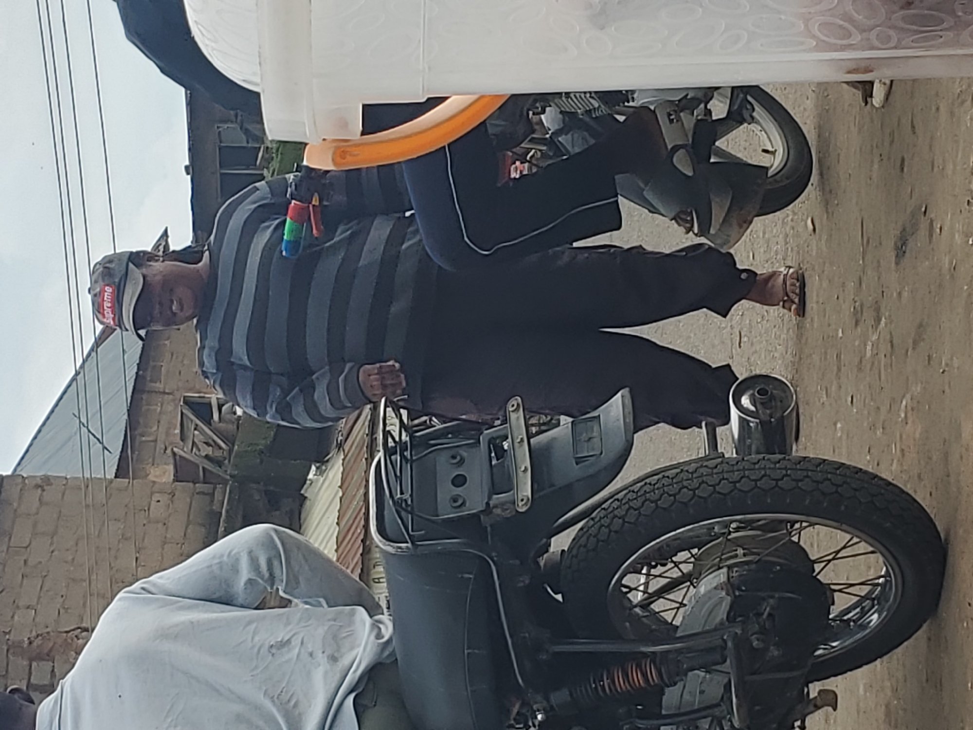 This image, taken in Gwong, Nigeria, depicts two people near a motorcycle. The primary subject, a person wearing a grey and black striped long-sleeved shirt, a grey cap with "Supreme" written on it, and dark pants, is holding a toy water gun. They are positioned on the right side of the frame, partially obscured by a large white container. The person is looking towards the left, and their posture suggests they might be interacting with the other individual or preparing to use the water gun.
The second person, wearing a light blue long-sleeved shirt, is visible in the lower left corner of the image, with their back to the camera. They appear to be sitting or leaning.
The scene is outdoors, with a dirt or gravel ground. In the background, there are buildings with corrugated metal roofs and brickwork, indicating a residential or semi-urban setting. Power lines are visible in the upper left portion of the image. The lighting suggests it is daytime, possibly overcast, as there are no strong shadows.
Notable details include the specific design of the water gun, with its red, blue, and green components, and the "Supreme" logo on the person's cap. The motorcycle itself is a prominent object, with visible wheels, an exhaust pipe, and part of the frame. The overall impression is one of casual activity in a local Nigerian environment.