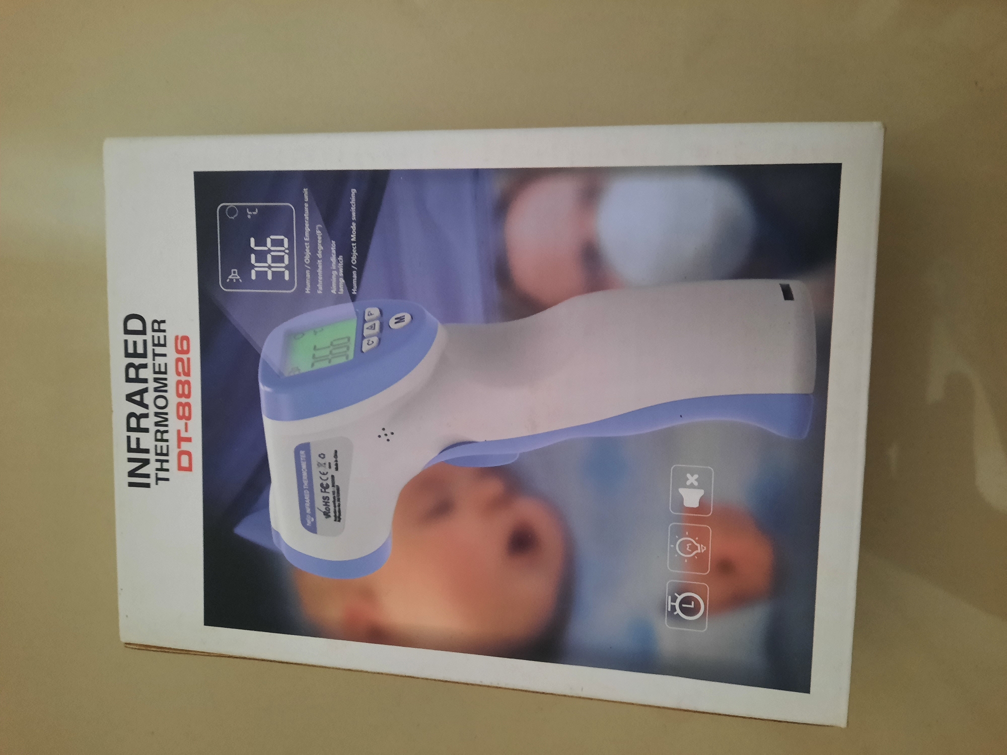The image displays the retail packaging of an infrared thermometer. The box is white with blue and red lettering. The front of the box features a picture of the thermometer, with a digital display showing "36.6°C". Text on the box indicates it is an "INFRARED THERMOMETER" with the model number "DT-8826". There are also icons representing a clock, a lightbulb, and a speaker with a cross through it, suggesting features like a timer, backlight, and mute function. The background of the box shows a blurred image of a baby and a person wearing a mask. The box itself is placed on a neutral, light-colored surface, likely a table or floor, with a plain wall in the background. The lighting is moderate and appears to be from an overhead source. The image does not contain any discernible elements that would allow for its location to be identified as Lhokseumawe, Indonesia.
