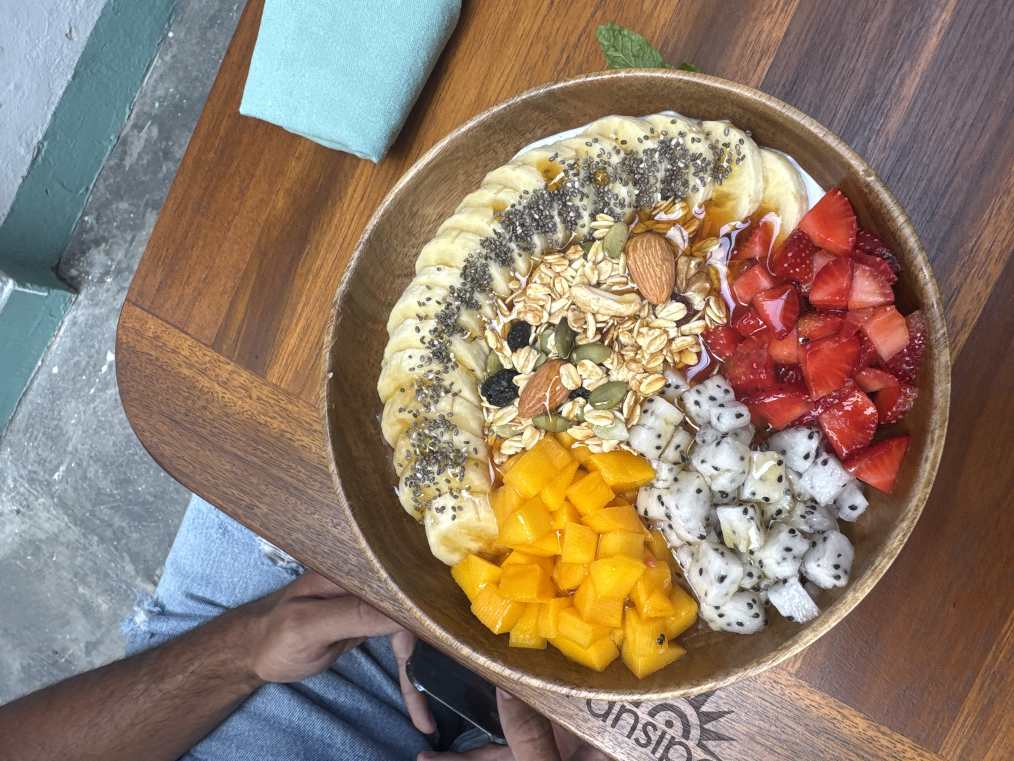 A high-angle, eye-level shot captures a wooden bowl filled with a smoothie bowl, likely photographed in Đà Nẵng, Vietnam. The bowl contains an array of fruits and toppings arranged in segments. Banana slices sprinkled with chia seeds line the top left, adjacent to a granola mix with nuts. To the right, diced strawberries gleam with a glossy sheen. Below the granola, cubed mango pieces sit next to cubed dragon fruit. A light blue cloth rests on the wooden table next to the bowl. A person wearing blue jeans is partially visible, holding a black object, possibly a phone, beneath the bowl. The wooden table has a logo at the bottom. The background includes a glimpse of a grey and blue wall and flooring. A sprig of mint garnishes the top of the bowl.