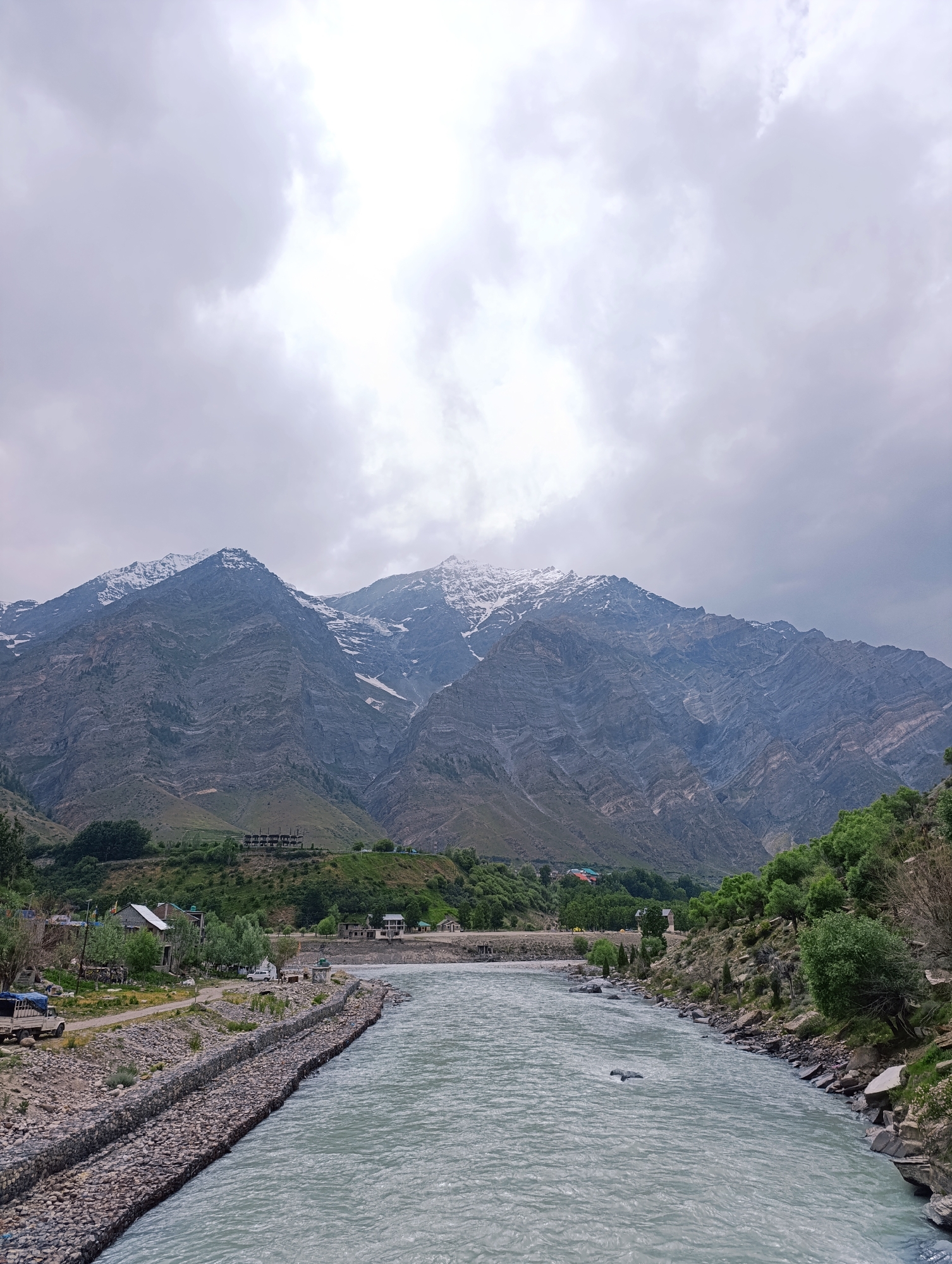 The image shows a wide, fast-flowing river in the foreground, with its milky blue-grey water rippling. The riverbanks are lined with stone-filled gabion walls on the left and a rocky, vegetated slope on the right. On the left bank, a dirt road runs alongside the river, with several small buildings and a parked truck. Beyond the road, the landscape rises to a series of green, terraced hills. In the background, imposing, jagged mountains dominate the scene. These mountains are a mix of dark grey rock with patches of snow on their upper reaches. The sky above is overcast with thick, grey clouds, obscuring any direct sunlight. The scene appears to be located in a mountainous region, consistent with the area around Tandi, India, known for its rivers and mountain ranges. There are no people visible in the image. The overall impression is one of a rugged, natural landscape under a dramatic sky.