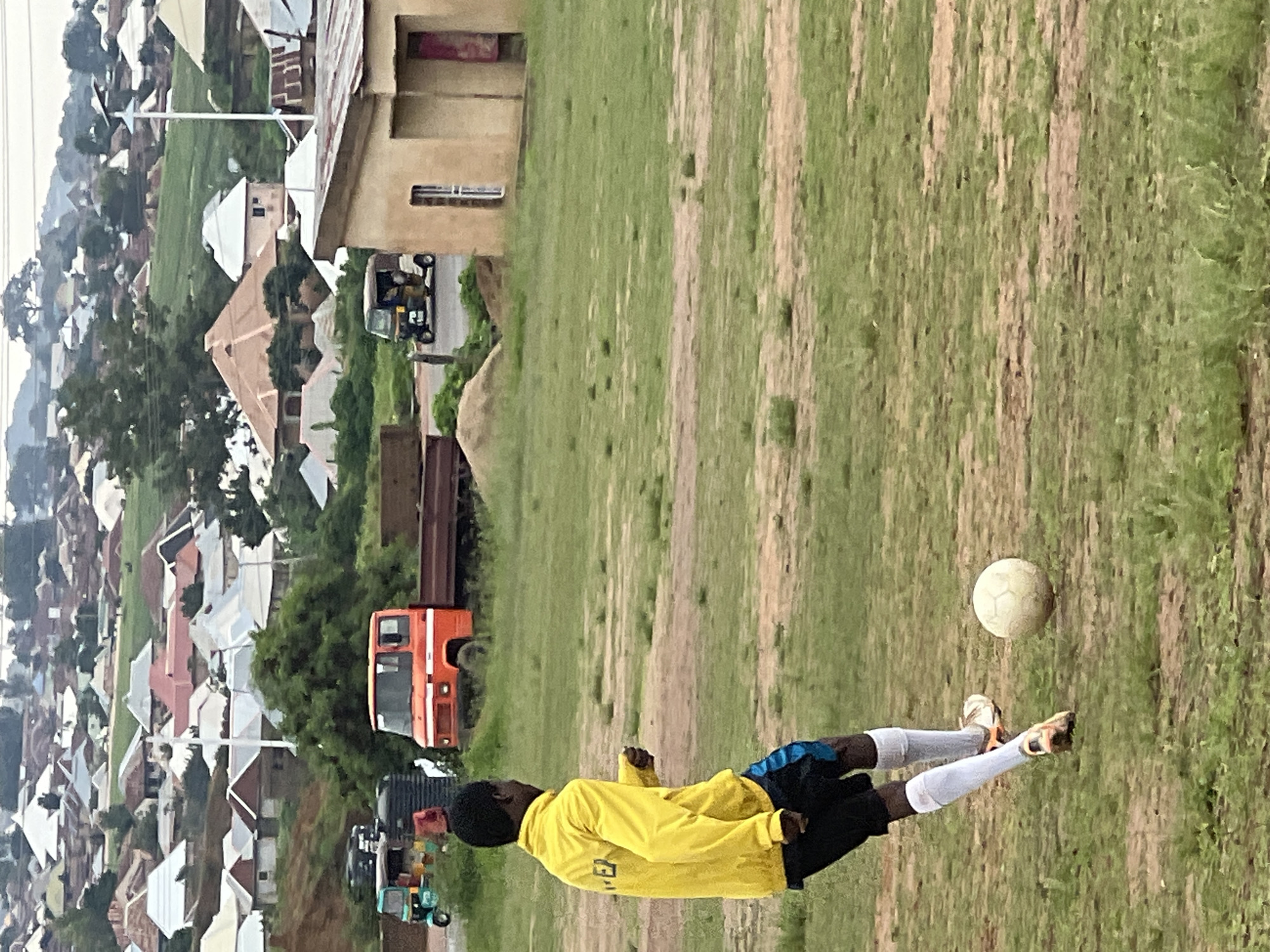 An outdoor, medium-range shot captures a young Black male in a yellow hooded jacket and black shorts playing soccer on a grassy field. He is kicking a white soccer ball with his right foot, his body angled towards the left of the frame. The man wears white knee-high socks and orange and white soccer cleats. In the background, a dusty, red truck is parked next to a pile of dirt. To the left of the truck, a green and blue auto-rickshaw is visible. Beyond the immediate area, a dense residential community with numerous houses, many with corrugated metal roofs, sprawls across rolling hills. The architecture suggests a semi-urban or rural setting. The sky is overcast, indicating a cloudy day. The overall environment appears to be the city of Jos, Nigeria, characterized by its hilly terrain and distinct housing styles.
