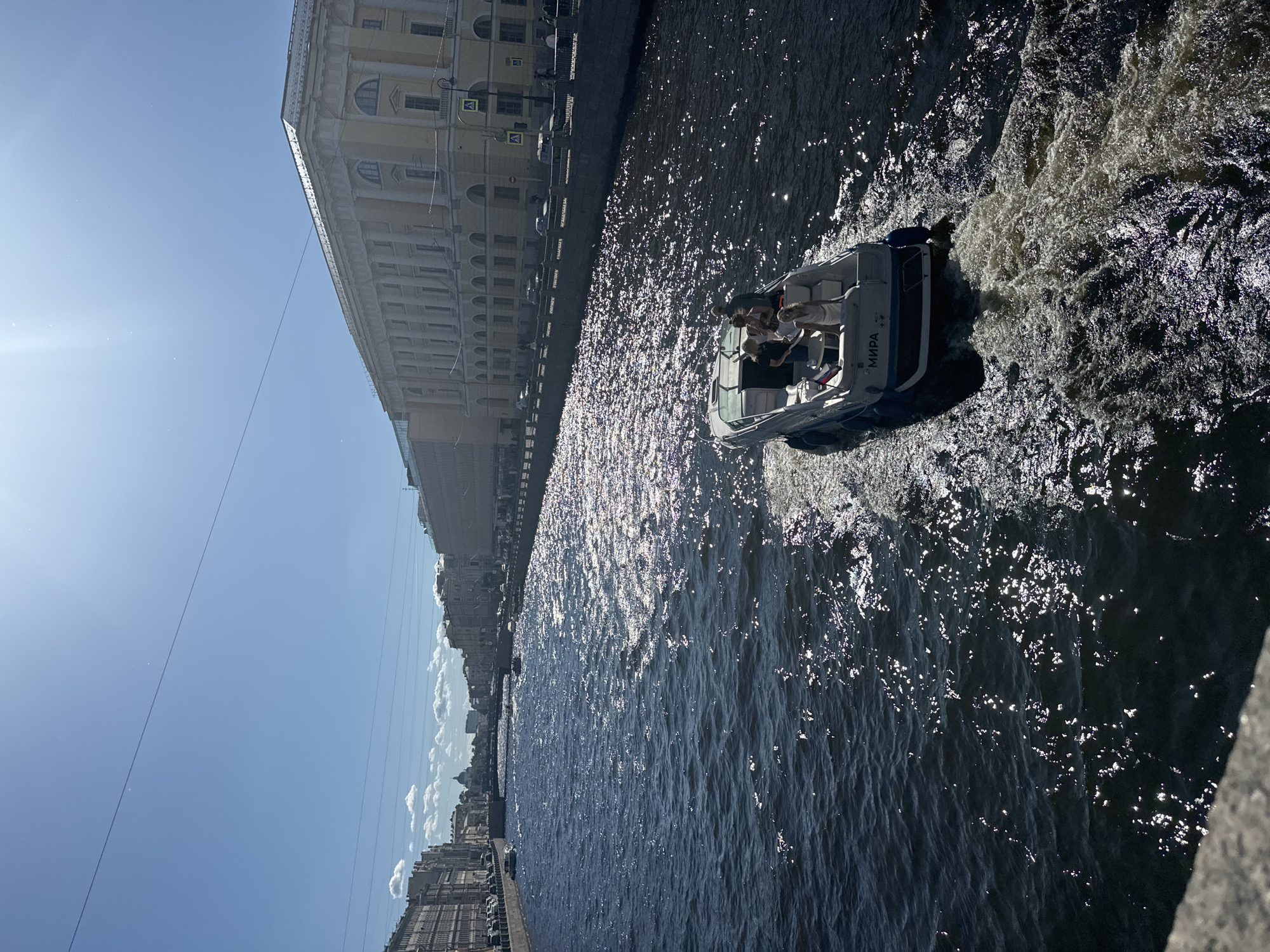 A motorboat, with the name "МИРА" visible on its stern, travels along a canal in Saint Petersburg, Russia. The boat carries several passengers, some standing and some seated. Sunlight glints off the dark water, creating a sparkling effect. On the right bank of the canal, a long, multi-story building with classical architecture lines the water. The building features numerous windows and a colonnaded facade. The canal is flanked by embankments, and further down, bridges can be seen spanning the water. The sky above is clear and blue, with a few scattered clouds and a visible power line. On the left bank, more buildings and trees line the canal, with cars visible parked along the street. The overall scene depicts a sunny day with recreational activity on the canal.