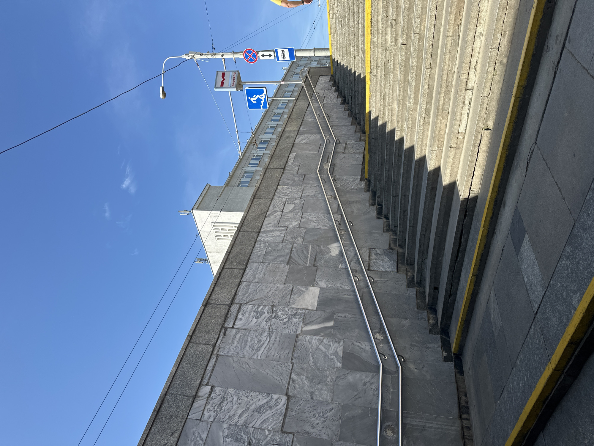 The image depicts an outdoor scene in Minsk, Belarus, featuring a set of concrete stairs leading upwards alongside a marble-clad wall. The stairs have yellow markings on their edges. Above the wall and stairs, a light-colored building with numerous windows is visible, situated under a clear blue sky. A lamppost stands near the stairs, adorned with several traffic and pedestrian signs, including a blue sign with a white pedestrian symbol and the Russian word "переход" (perekhod), meaning "crossing." Other signs on the lamppost include a red circular sign with a diagonal line, indicating prohibition, and a square sign with a bus icon and an arrow. Overhead power lines crisscross the sky. The overall scene suggests a public area, possibly an underpass or a pedestrian walkway in an urban environment.