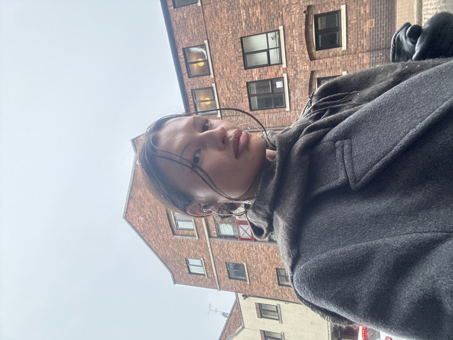 A female subject, seen from the chest up, is positioned outdoors on an overcast day. She has dark hair, with strands framing her face, and wears a light grey wireless earbud in her left ear, along with large silver hoop earrings. She is dressed in a dark grey heavy coat and a charcoal-colored scarf with visible fringed edges. The subject gazes directly forward. The background consists of brick buildings; a multi-story red-brick structure with several windows, some illuminated from within, is prominent behind her. To the left, a lighter brick building with a red architectural accent and an antenna is partially visible. The urban architectural style and overcast conditions are characteristic of a city setting in London, United Kingdom. No specific actions or interactions are apparent.
