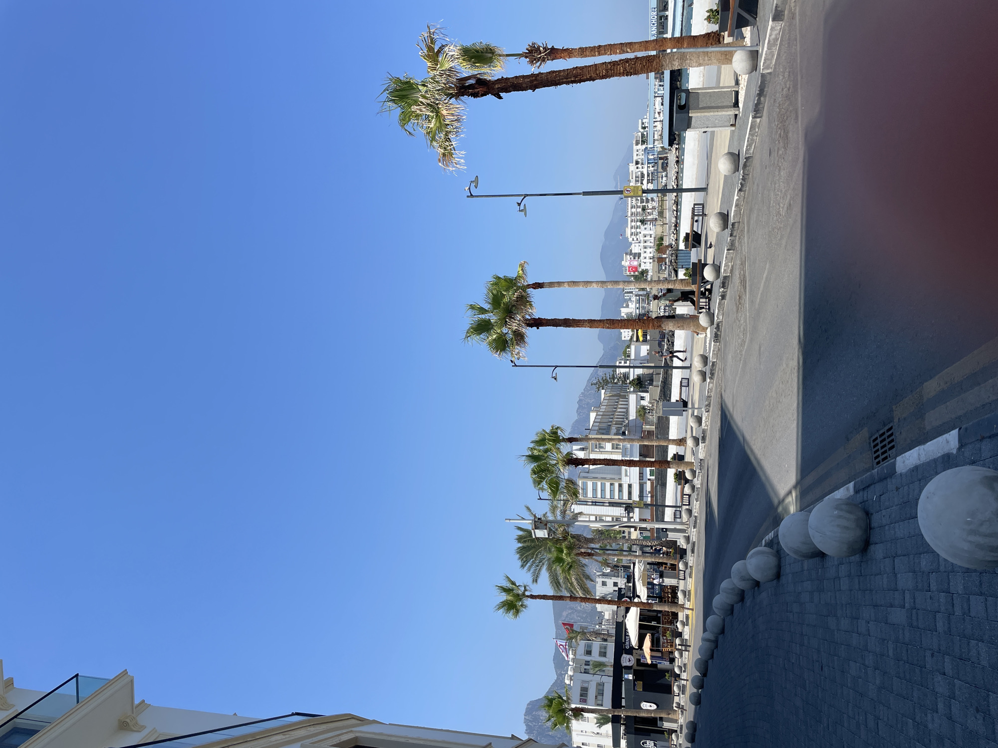 A long shot captures a coastal promenade in Kyrenia, Northern Cyprus, under a clear blue sky. The scene is dominated by a row of tall palm trees lining a wide, paved walkway adjacent to a street. The walkway features numerous large, spherical concrete bollards. Beyond the palm trees, a white, modern building with balconies and glass railings is visible on the left, with the clear blue sky extending above it. Across the street, a line of white and light-colored buildings, some appearing to be hotels or apartments, stretches into the distance, with mountains forming a backdrop. Several flags are visible, including what appears to be the flag of Northern Cyprus. People are sparsely scattered, with a few individuals walking along the promenade and seated on benches. A few outdoor cafe tables with umbrellas are also present. A trash receptacle stands near the right edge of the image. The sea is visible in the background, with a boat docked in the harbor.