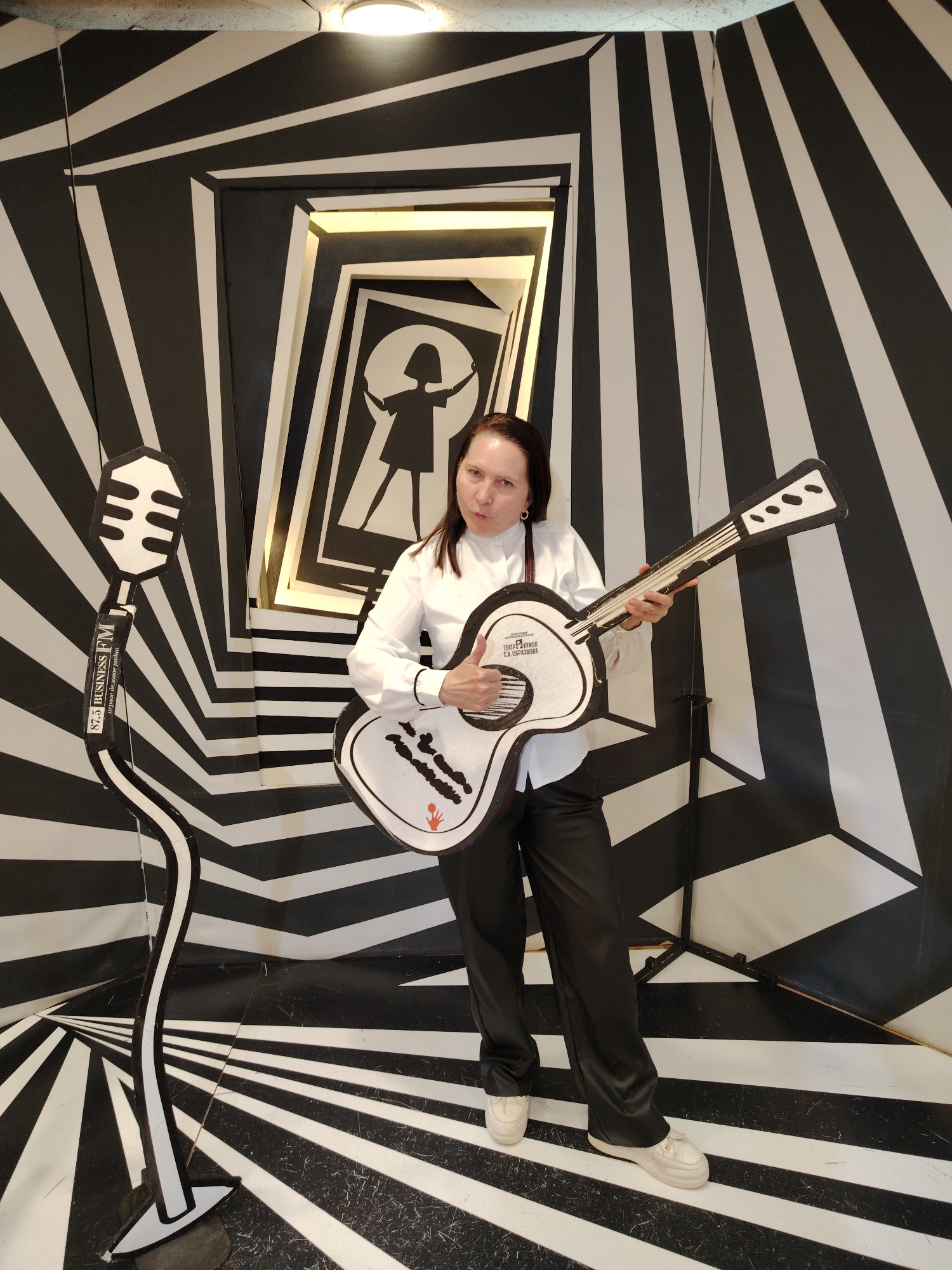 A full shot captures a middle-aged woman indoors, posing with props in a visually striking, optical illusion-themed setup. The backdrop features a black and white striped pattern creating a tunnel effect. The woman is dressed in a white button-down shirt and black pants, and she is holding a large, cartoonish white guitar. To her left, a similarly styled microphone prop stands. The location is the Central Children's Store on Lubyanka Square in Moscow, Russia.