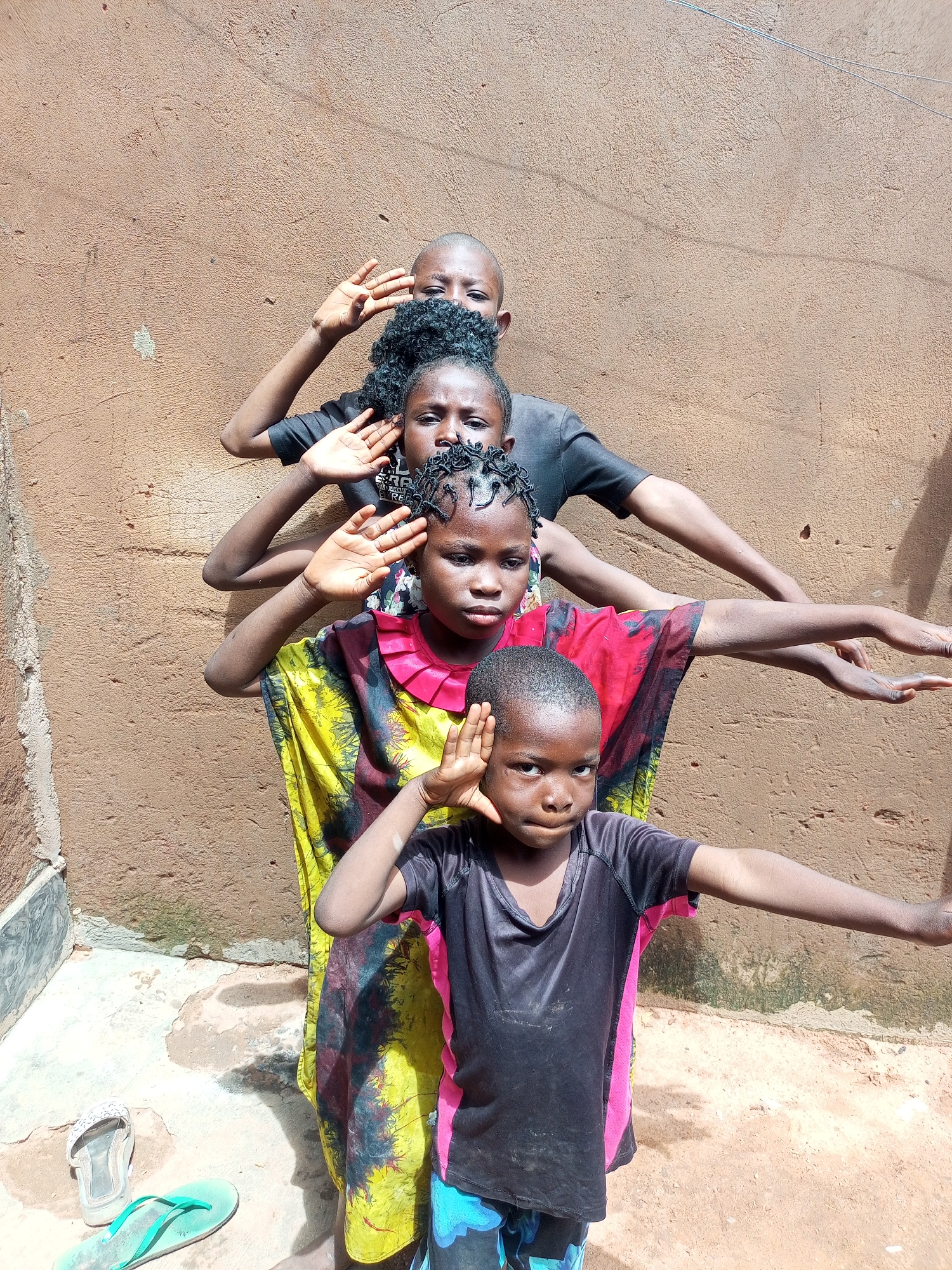 Four children are posing in a line against a textured, brown wall. From front to back, they are: a young boy wearing a black and pink t-shirt and blue patterned shorts, a girl in a yellow and red patterned dress, another girl with curly hair wearing a black t-shirt, and a child with a shaved head in the back, also in a black t-shirt. All four children have their arms extended outwards, with their hands open and held at head height, appearing to be in a synchronized pose. They are standing on a concrete ground with some sand and debris. To the left of the frame, a pair of light-colored flip-flops and a pair of green flip-flops are visible on the ground. The scene appears to be outdoors in an urban setting, possibly in Gombe, Nigeria. The lighting suggests it is daytime, with shadows cast by the children on the wall.
