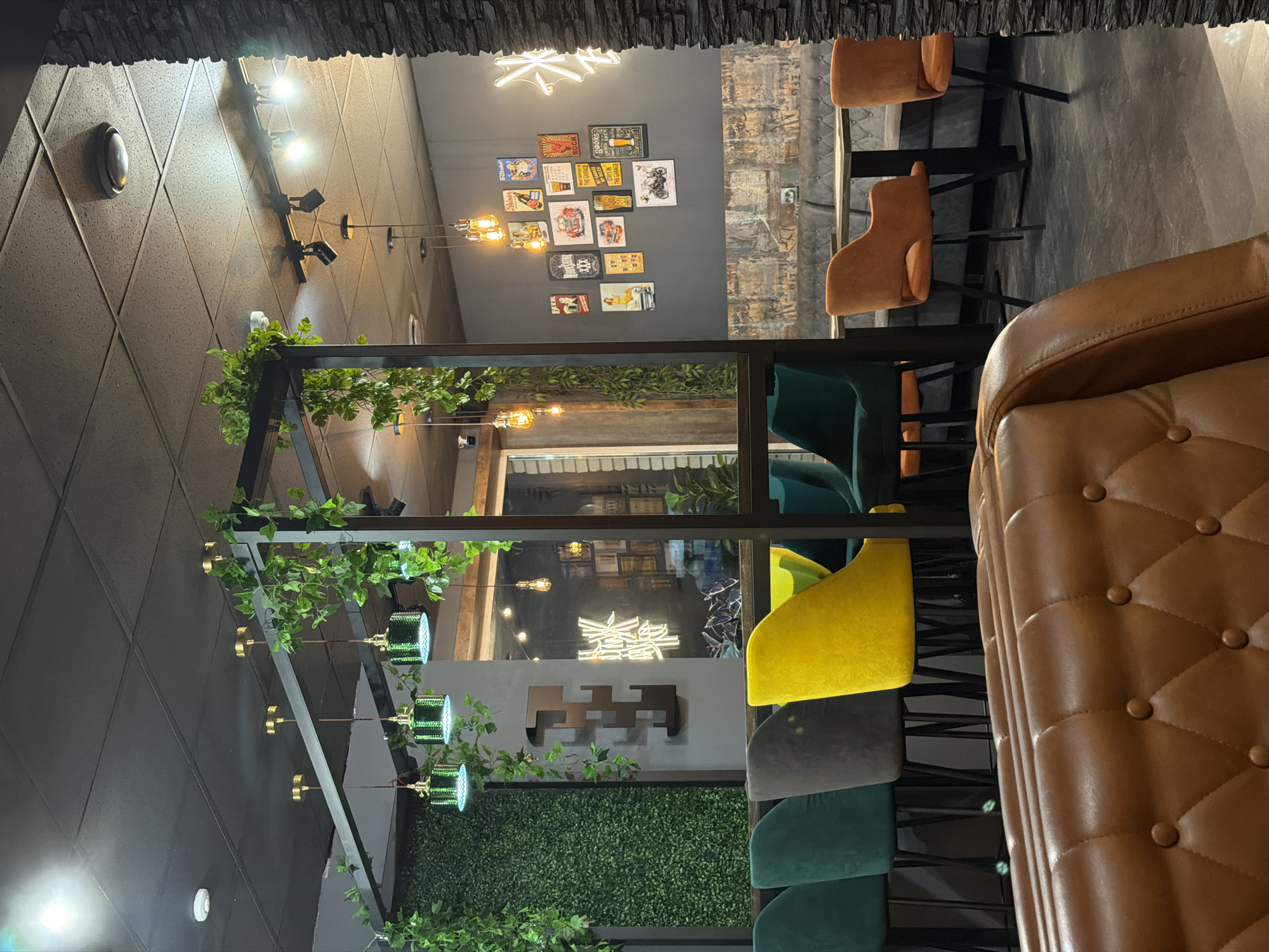 The image shows the interior of a restaurant or bar. The scene features a black metal structure adorned with foliage, which acts as a visual partition. Behind it, there is a seating area with various colored chairs (yellow, green, gray). A brown leather booth is visible in the foreground. The restaurant's décor includes lights, pictures on the wall, and a decorative panel with Chinese characters. The city name, Sarapul, Russia, is not discernible in the image.