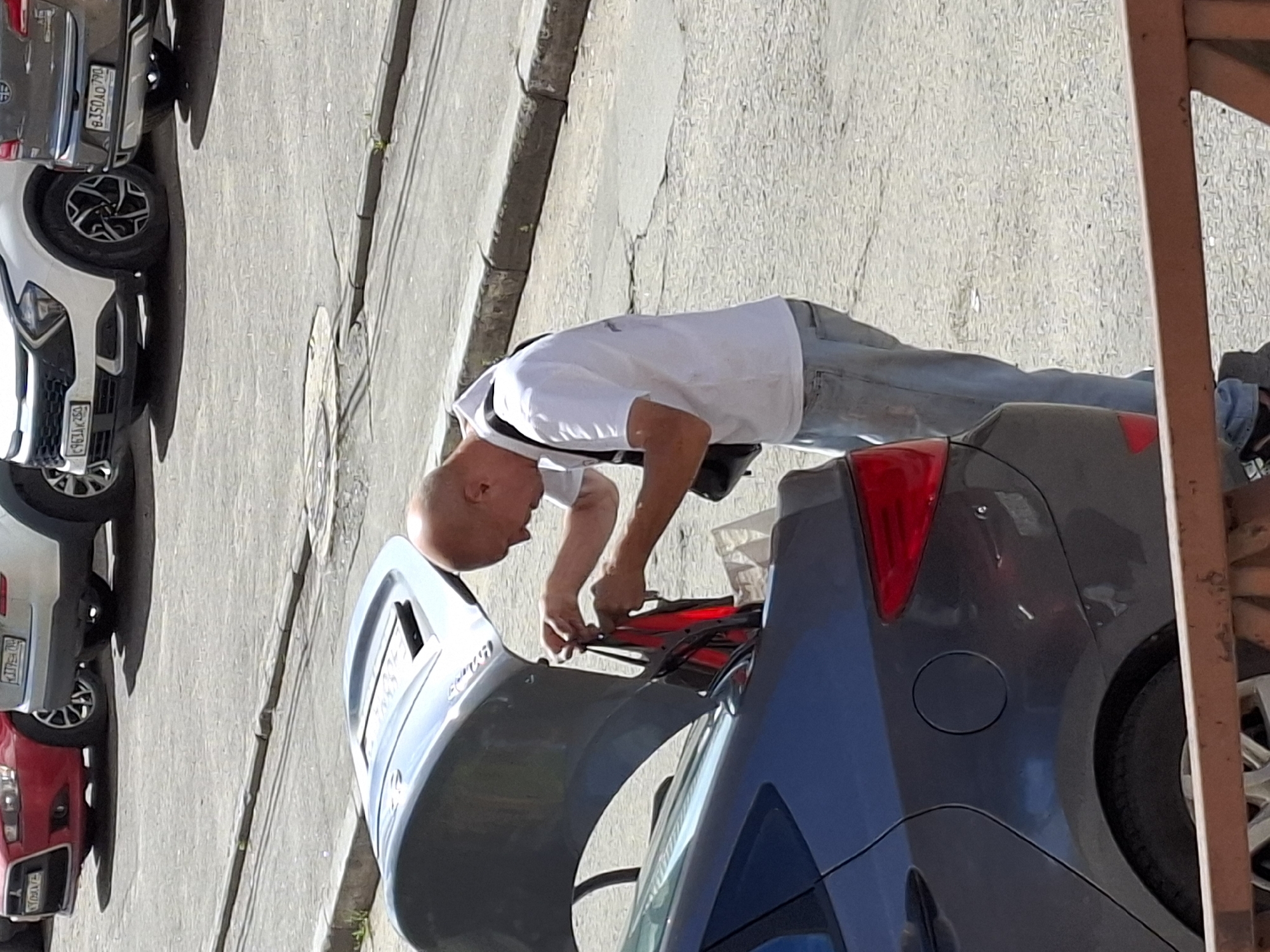 In a street scene, a bald man wearing a white t-shirt and blue jeans is seen from the side, standing next to the open trunk of a dark gray car. He is actively engaged in an action, appearing to be handling some red objects, possibly clothing or bags, that are within the trunk. A camera is slung around his neck. The car's trunk is elevated, revealing its interior. Other cars are parked in the background, suggesting a public parking area or street. The ground is paved and appears to be asphalt or similar material, with some cracks and curbs visible. The lighting indicates it is daytime, with shadows cast on the ground.