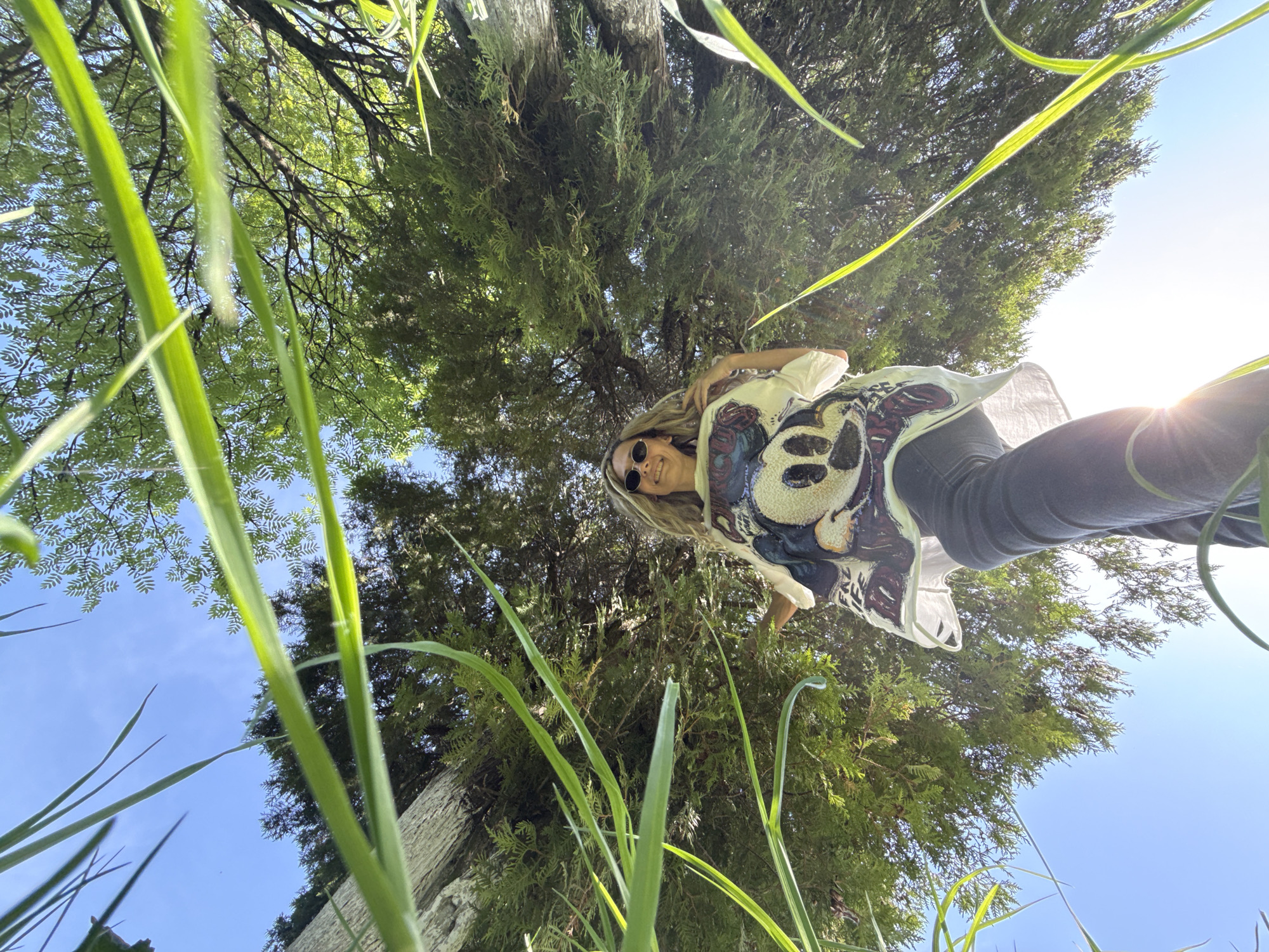 The photo is taken from a low angle, looking up at a person standing above. The person is wearing sunglasses, a light-colored patterned shirt, and dark pants. They are positioned against a backdrop of lush green foliage, including grass in the foreground and trees filling the background. The sky is visible in the upper left and right corners, suggesting a bright day. The image was not taken in Rusanovo, Russia.