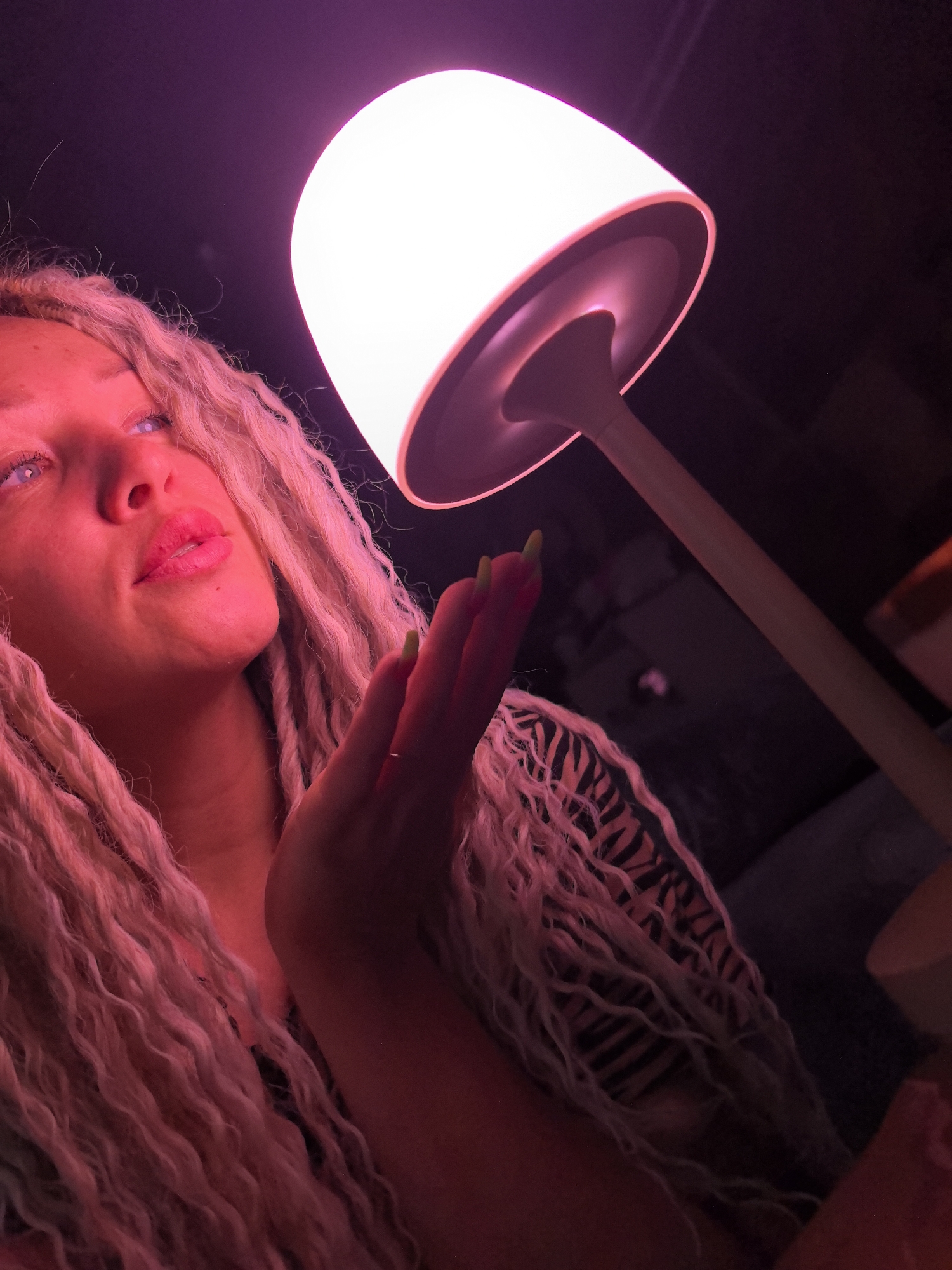 A woman with long, blonde, curly hair is angled upwards, illuminated by a pinkish-white light emanating from a small table lamp to her right. She has fair skin, blue eyes, and pink lips. Her hand is raised towards the light. The woman is wearing a black and tan striped top. The background is dark, but what appears to be a sofa can be seen behind the lamp. The location, Noviy, Russia, cannot be determined from the image.