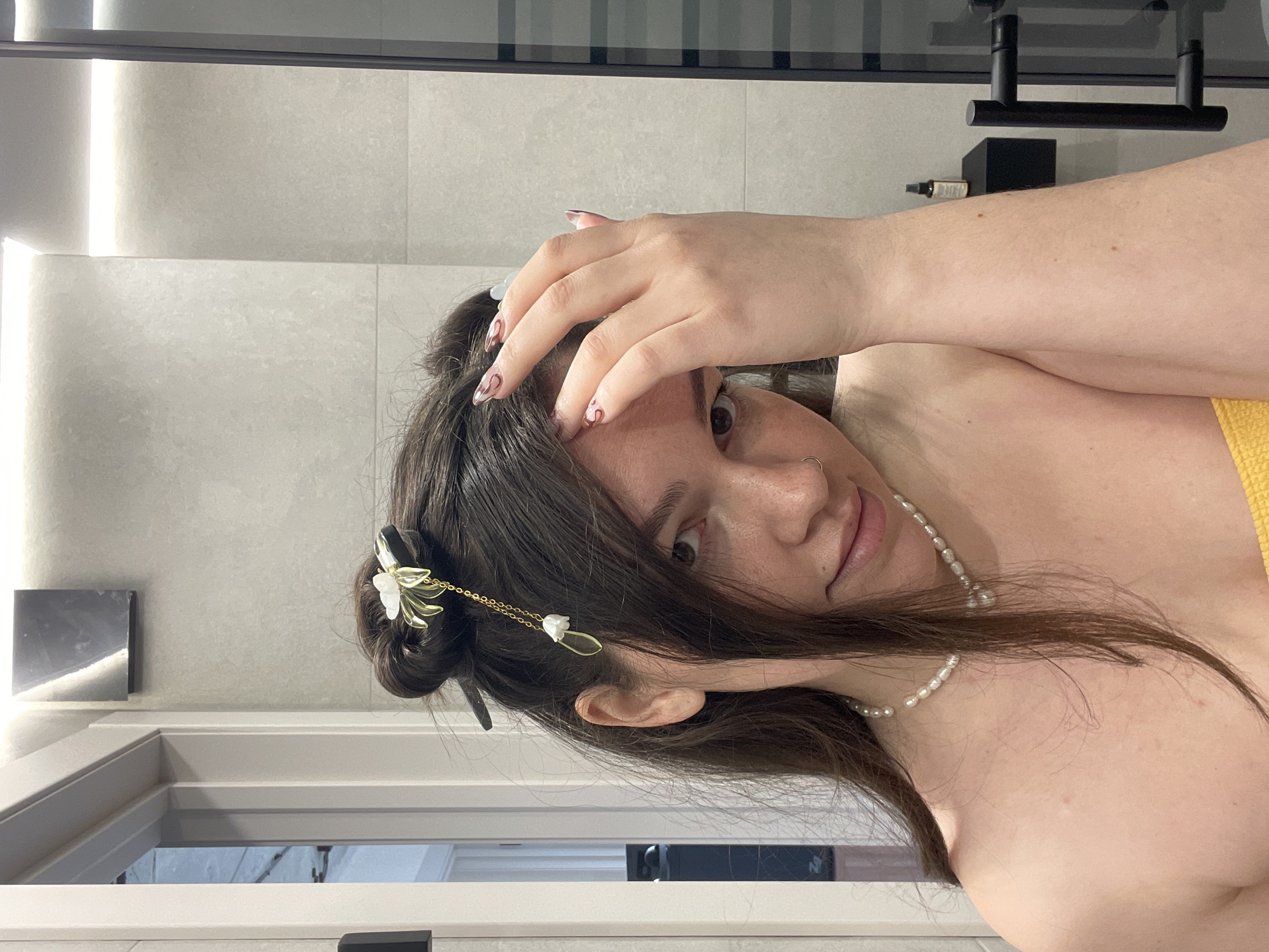 A young woman is captured in a close-up, rotated portrait, likely taken in a modern bathroom or shower setting. Her dark brown hair is styled, with some strands pulled back and adorned with an ornate, gold-toned hair clip featuring white floral details and delicate dangling chains with small white beads. She wears a pearl necklace around her neck and a silver hoop nose piercing. A yellow garment, possibly a towel or top, is visible on her right shoulder.
The woman holds her right hand to her forehead, partially obscuring the upper left side of her face. Her fingernails are long and almond-shaped, painted with a clear base and subtle dark, abstract swirl designs. Her right eye is wide open and looks directly towards the viewer, conveying a direct or slightly curious gaze. Her lips are slightly parted.
The background consists of light grey tiled walls. To the left, a vertical strip of bright light suggests integrated lighting. On the right, black bathroom fixtures, possibly a shower head or controls, and a small dark bottle on a ledge are visible. A reflective surface, likely a mirror or glass partition, is also seen in the lower left portion of the image. The overall lighting is bright and even. There is no visible text.