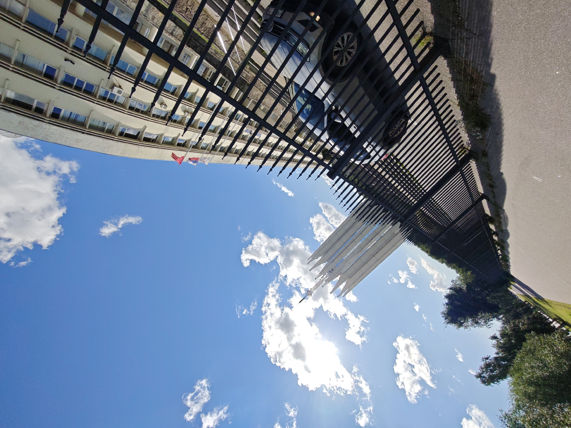 This image depicts an outdoor scene with a black metal fence in the foreground, separating a paved walkway from a grassy area and a building in the background. The fence has a pattern of vertical bars topped with pointed finials. A light-colored car is parked behind the fence, partially visible. The building is a multi-story structure with many windows and what appear to be air conditioning units mounted on its facade. Several flags, including what looks like the Russian flag, are flying from the top of the building. Above the building, a tall, modern, white spire-like structure rises into a bright blue sky with scattered white clouds. The sun is visible, casting shadows and creating a glare. Trees are present to the left of the walkway, with green grass and some shrubbery visible between the fence and the building. The angle of the shot suggests the photographer is looking upwards and to the right. The scene appears to be in Saint Petersburg, Russia, given the presence of the Russian flag and the architectural style.
