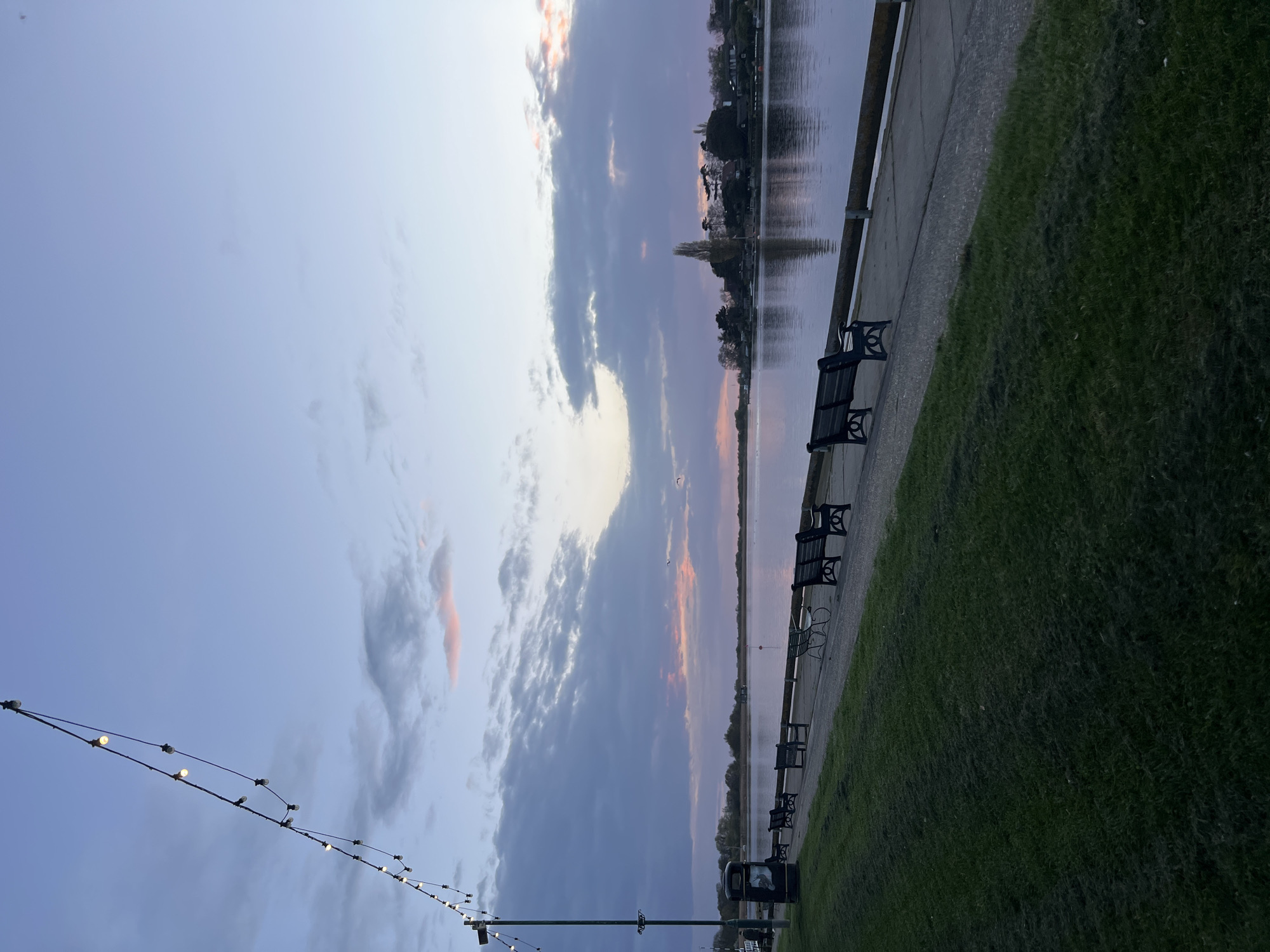 This image depicts a waterfront park scene at dusk in East Suffolk, United Kingdom. A series of black benches line a concrete walkway adjacent to a body of water reflecting the sky. Lush green grass extends away from the walkway. Strings of lights are visible on poles along the side. The sky is a mix of blue, gray, and pink hues with scattered clouds.