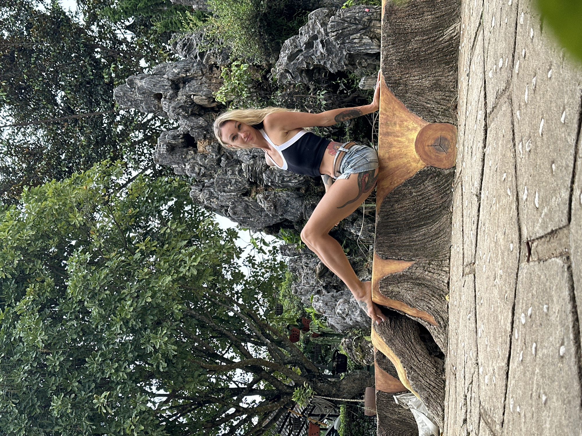 A woman with blonde hair is posing outdoors. She is wearing a black tank top with white trim and distressed denim shorts. She has tattoos on her arms and legs. She is leaning against a large, carved wooden structure that resembles a tree trunk. The carving is detailed with bark texture and a lighter wood tone in the center. To her left, there are rocky formations with lush green trees and foliage. The background suggests a natural, possibly tropical, environment. The scene appears to be set during the daytime with overcast or diffused lighting. The woman is looking towards the camera with a slight smile, suggesting a relaxed or playful mood. The ground in the foreground is paved with concrete stones. There is no visible text in the image. The context of Thôn 2, Vietnam, suggests a Southeast Asian setting.