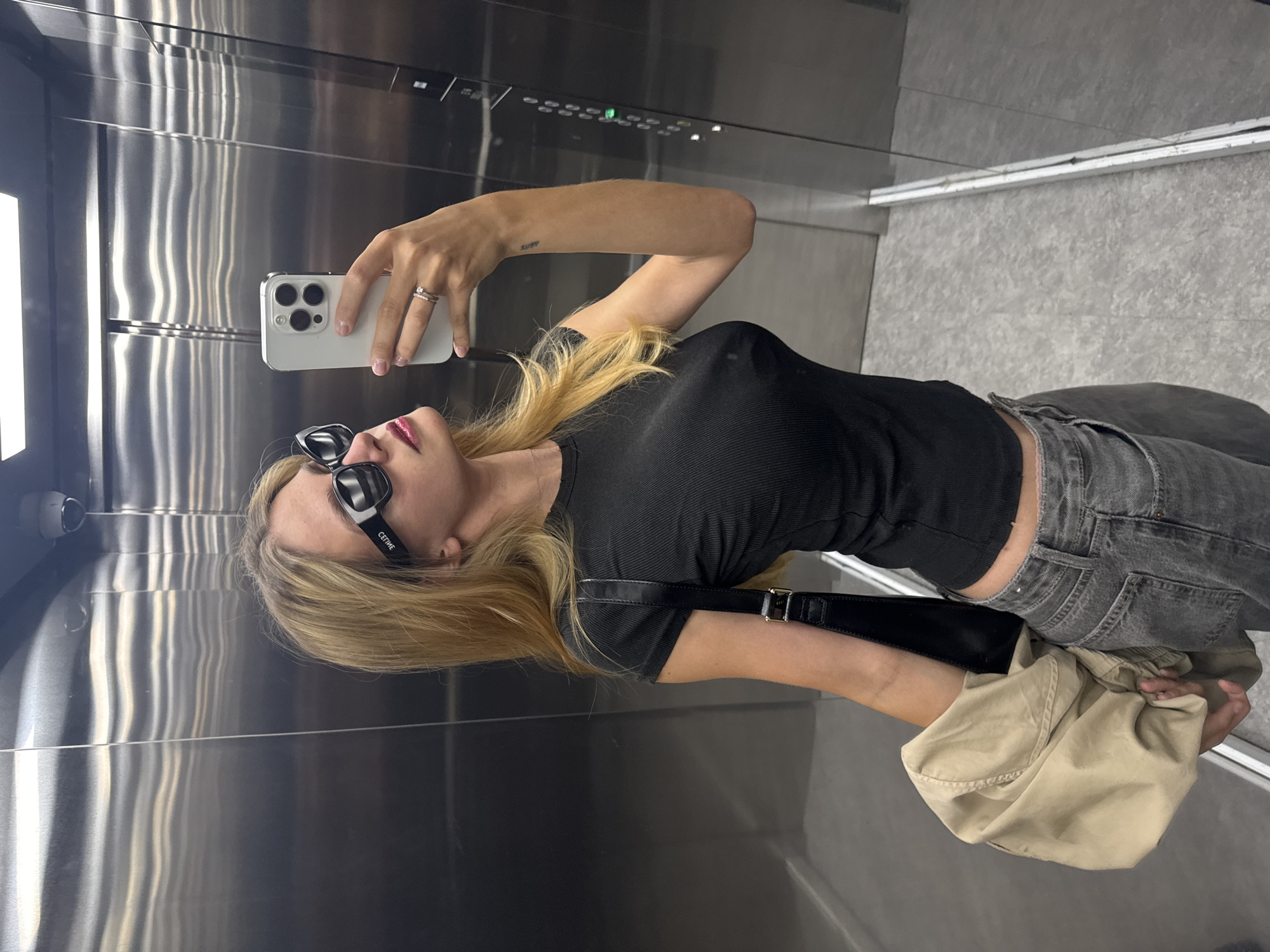 A medium shot depicts a fair-skinned person in an elevator taking a selfie with a white smartphone. They are wearing sunglasses, a black, cropped top, and gray jeans. A black purse hangs from their shoulder, and they are holding a beige jacket or bag. The elevator has stainless steel walls with buttons and indicators at the top. The floor is gray. The city and state are unknown.