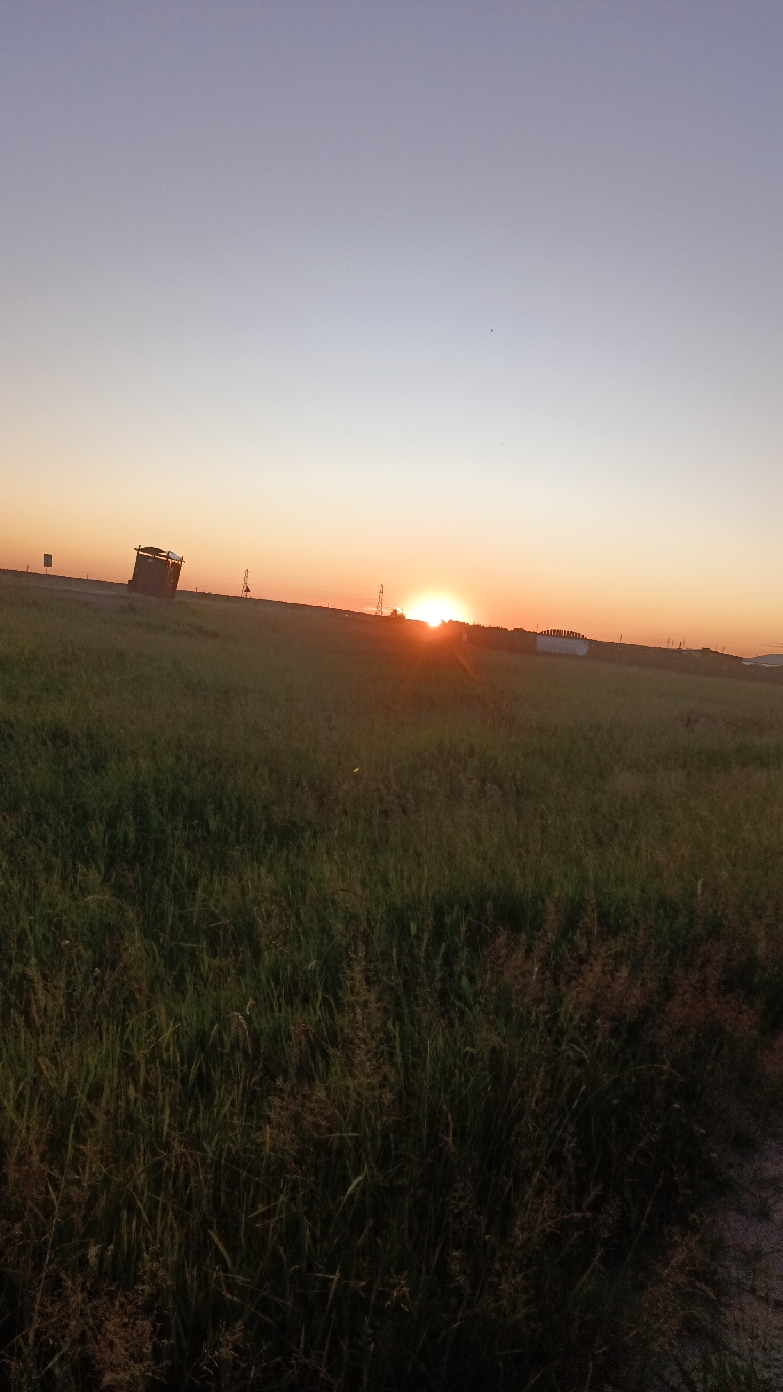 The image depicts a rural landscape at sunset. The foreground is dominated by tall, green grass, with some dried stalks mixed in. In the mid-ground, a brown, rectangular structure, possibly a small building or shelter, is visible on a slight incline. Several utility poles with wires are scattered across the horizon. The setting sun is a bright, orange orb just above the horizon, casting a warm glow across the sky, which transitions from a soft orange near the sun to a pale blue at the top of the frame. The overall impression is one of open space and the end of a day in a natural setting. This image was taken in the city of Angarsk, Russia.