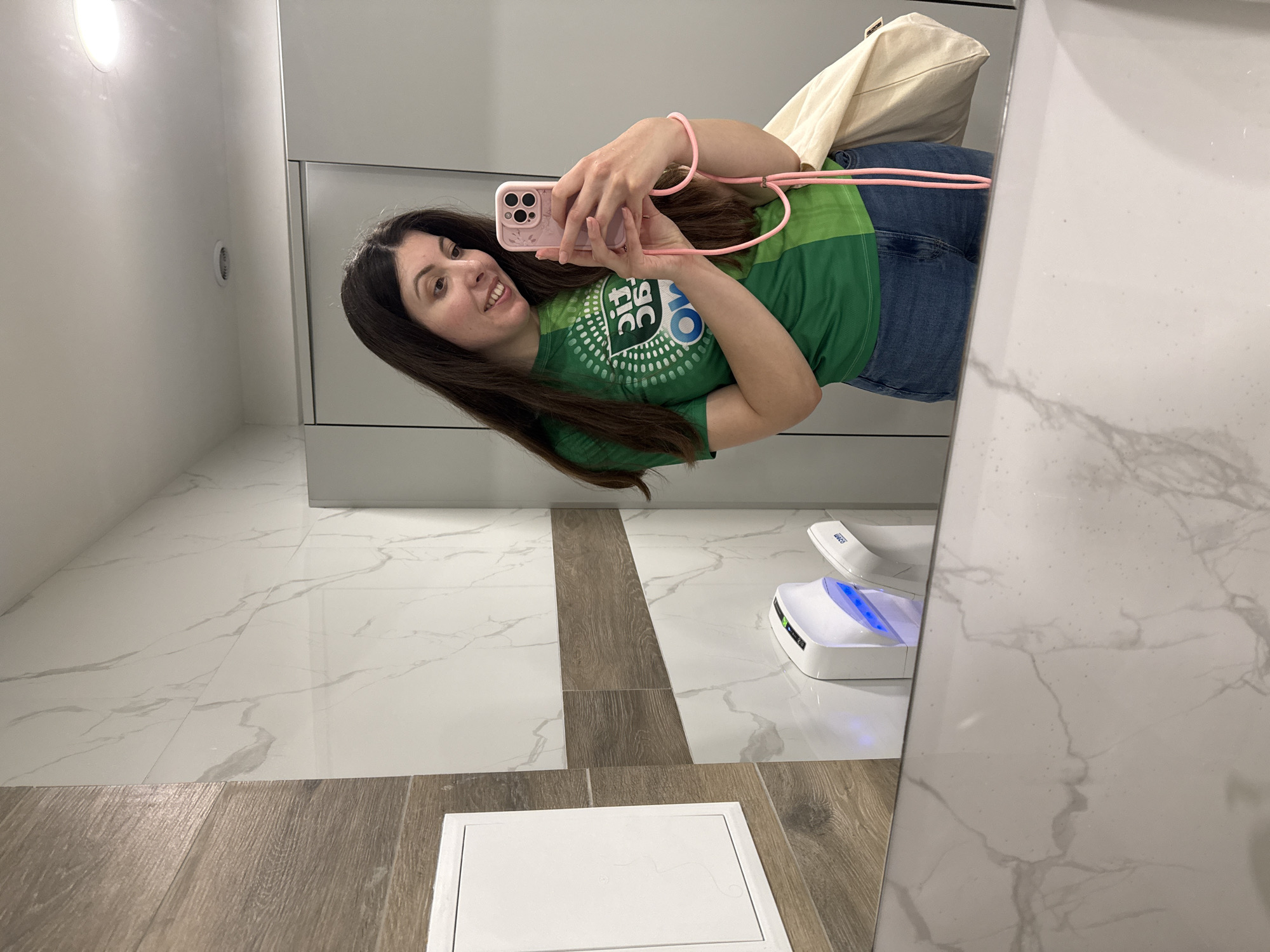 The image shows a woman taking a selfie in a bathroom mirror. She is wearing a green t-shirt, jeans, and a pink phone strap. She is holding a pink iPhone. Behind her is a toilet seat sanitizer on the floor and a beige tote bag. The floor is tiled with marble and wood-look tiles. The wall is white. The location is a bathroom, likely in a commercial building, city of Unknown, Unknown.
