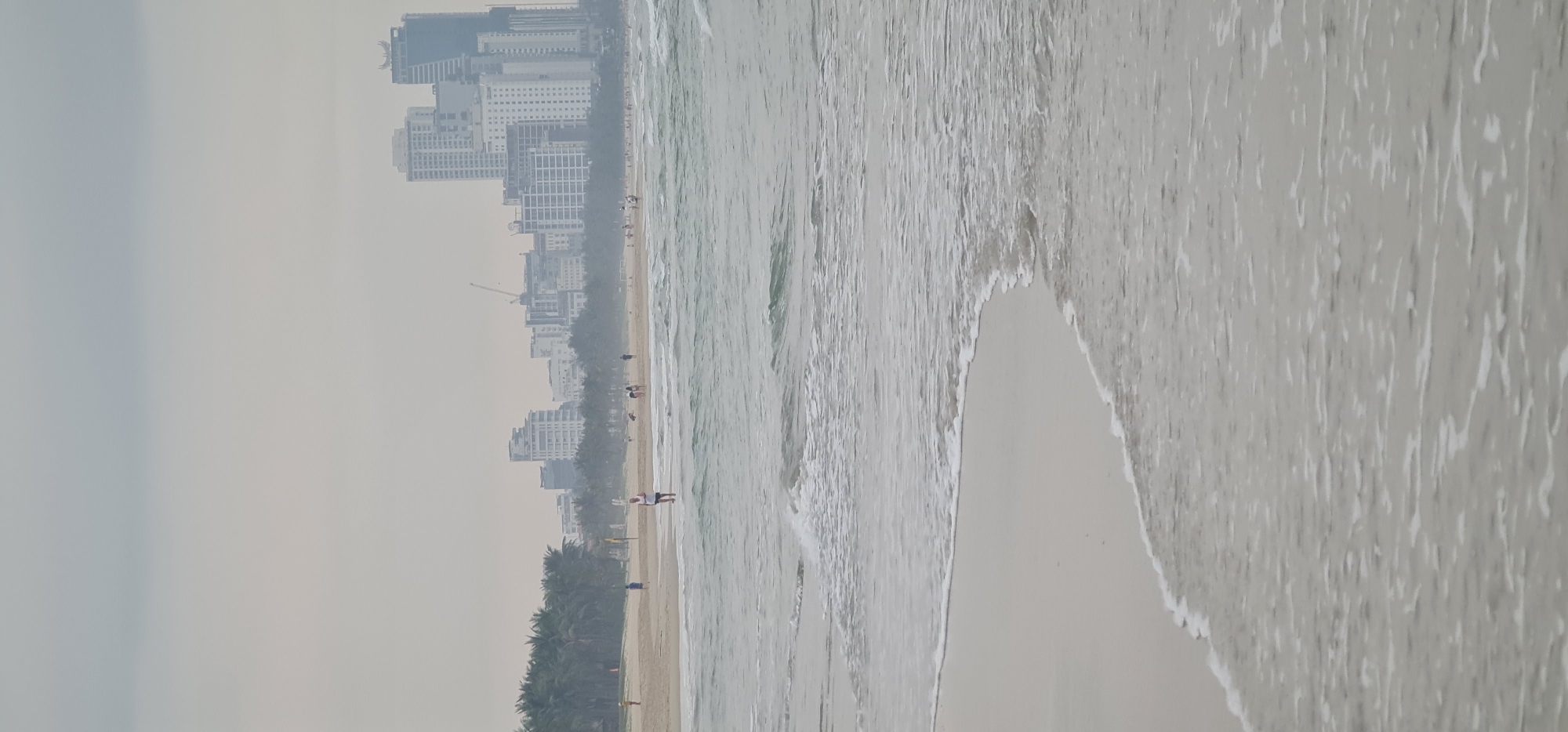 The image shows a coastal scene in Đà Nẵng, Vietnam. The foreground consists of a sandy beach with incoming waves. In the midground, the shoreline stretches out, lined with palm trees and a few scattered people. Tall buildings are visible in the hazy background.