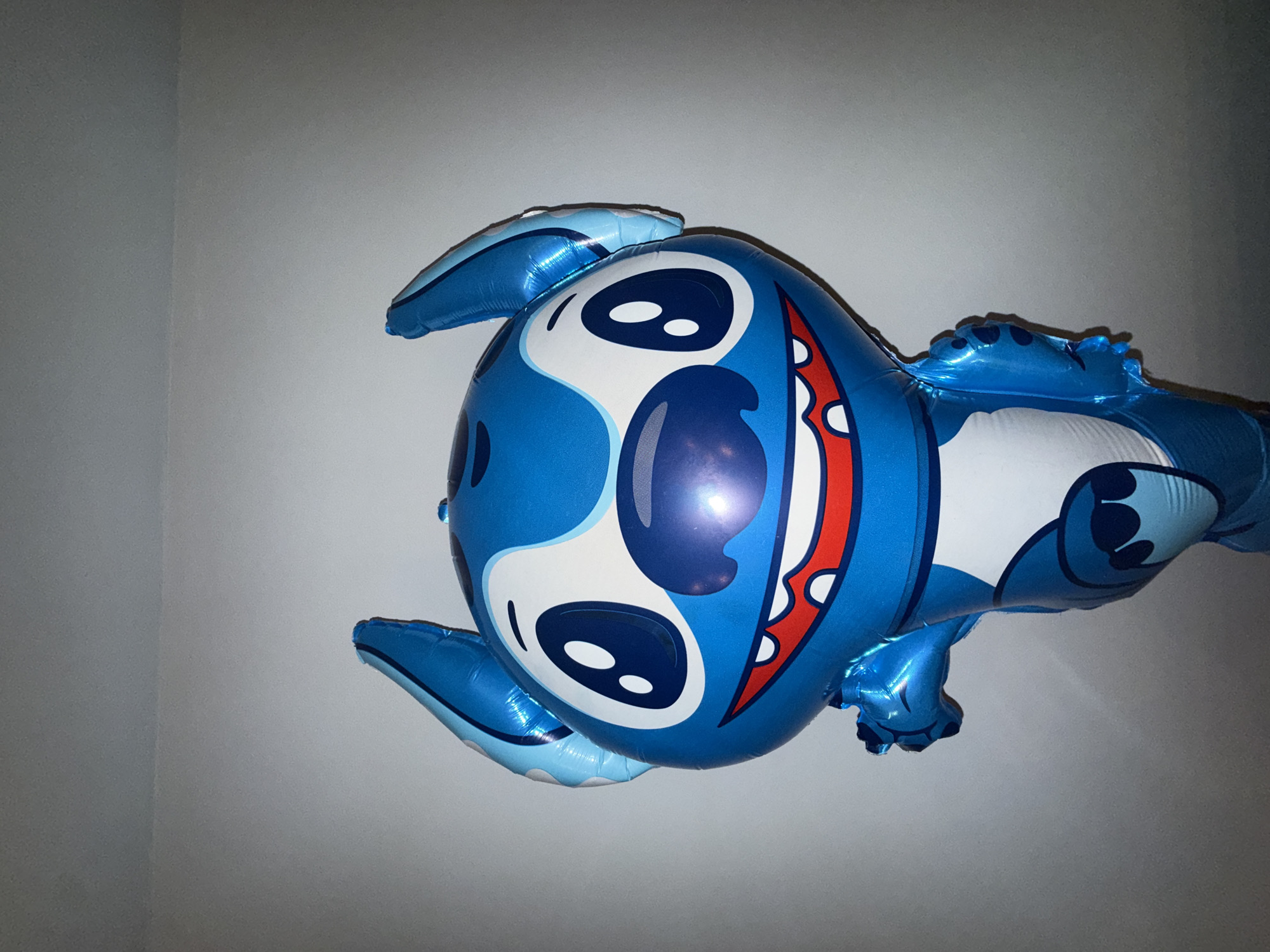 A close-up shot shows a blue Stitch-shaped foil balloon against a gray background. The balloon is inflated and displays Stitch's facial features, including his eyes, mouth, and ears. There is no visible indication that the image was taken in Blagoevgrad, Bulgaria. The background is plain and lacks any contextual information about a specific location.