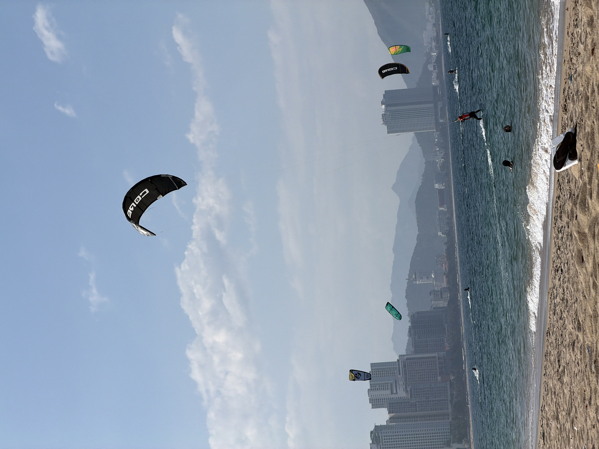 The image depicts a daytime scene on a sandy beach in Nha Trang Ward, Vietnam, with ocean activity and a city skyline backed by mountains.
The upper portion of the frame is dominated by a light blue sky with scattered white cumulus clouds. Centered prominently in the upper mid-ground is a large black kitesurfing kite with the word "CORE" visible in white, inverted text. To the left, a smaller yellow and black kite is partially visible. Further to the right, another small teal-colored kite is airborne. In the far right mid-ground, two additional kites are visible: one black with "CORE" branding, and an adjacent green and yellow kite. Thin lines extend from the kites towards the water, indicating connection to kitesurfers.
The middle ground features the deep blue-green ocean with moderate wave activity. A kitesurfer wearing an orange or red top is actively riding a board, emerging from the water in a probable jump or maneuver, with control lines visible. Further back in the water, another individual on a kitesurfing board is visible to the left of the main rider. Two more kitesurfers are active further in the background to the right. Closer to the foreground, two individuals are partially submerged in the water near the breaking waves, facing away from the camera, with their heads and shoulders visible.
The background reveals a densely built cityscape composed of numerous multi-story buildings, likely high-rise hotels or apartments, stretching along the coastline. Behind the city, a range of hazy, blue-tinted mountains forms the distant horizon.
The immediate foreground consists of light brown sand, characteristic of a beach. Gentle waves are breaking at the water's edge. Resting on the sand in the lower foreground, slightly right of center, is a white object, possibly a plastic bag or fabric, with a pair of black flip-flops placed on top of it.