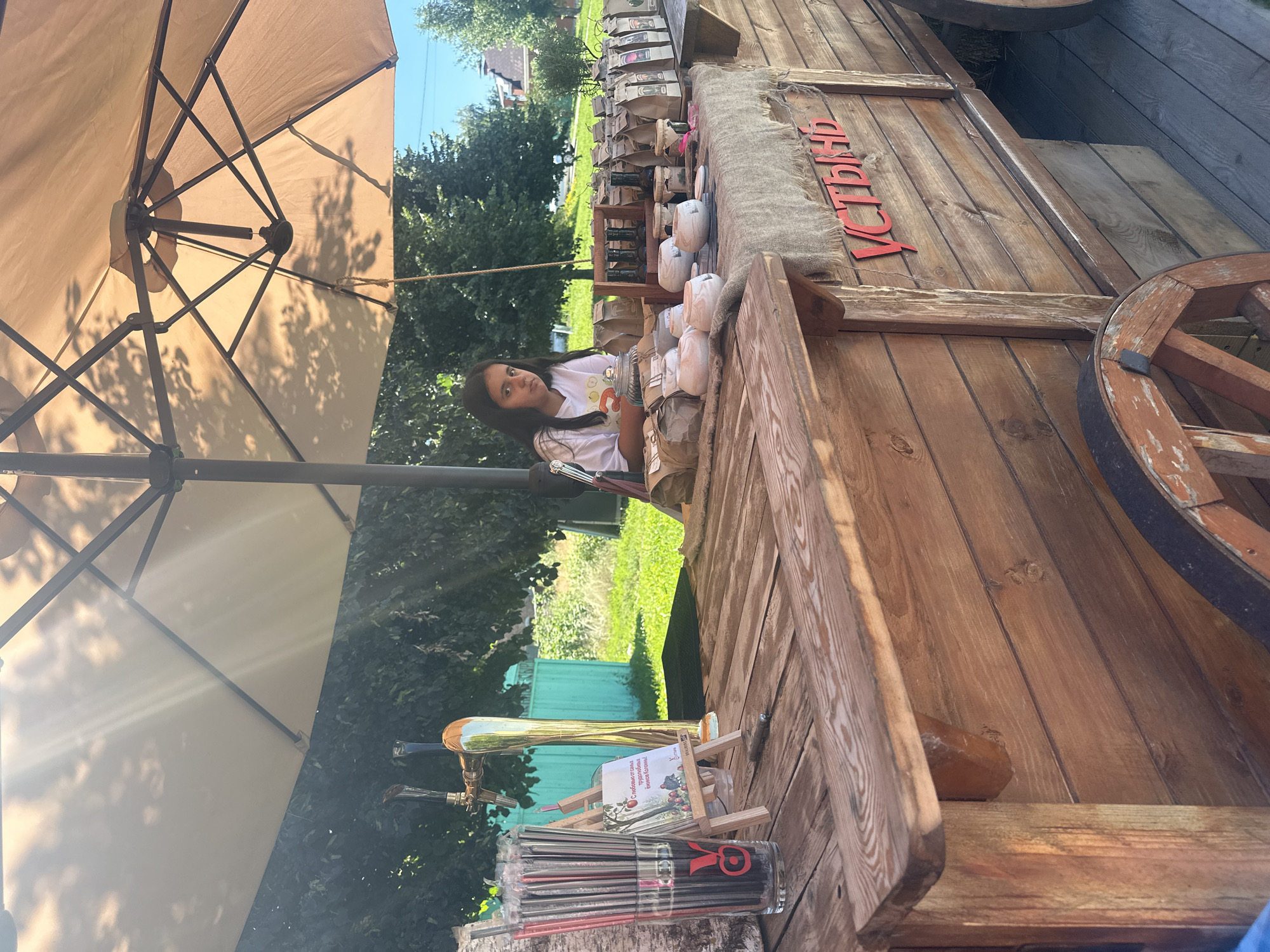 A young girl, likely of South Asian descent, sits behind a wooden market stall under a large beige umbrella. The stall is laden with various items, including bags of what appears to be tea or coffee, small ceramic bowls, and bottles. A beer tap is visible to the left of the girl, alongside a glass containing numerous straws. A small easel with a picture of an apple and text in Russian stands near the straws. The market stall is decorated with the Cyrillic word "УСТЬЯНЬ" in red letters. The background shows greenery and trees, suggesting an outdoor setting in a park or a market area. The overall impression is of a small, local business operating outdoors, likely in the city of Kolomna, Russia, given the Cyrillic signage.