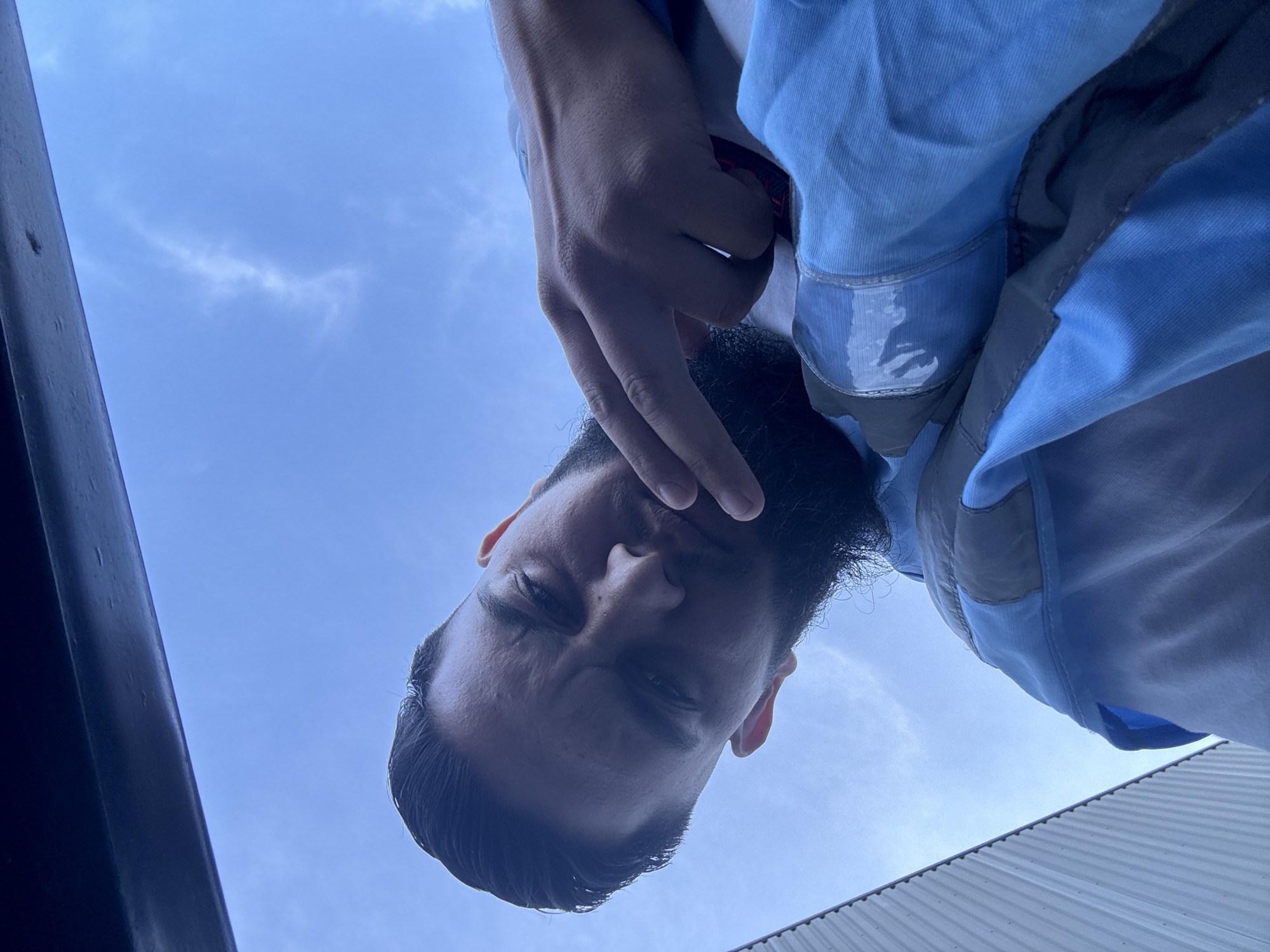 A low-angle, close-up, eye-level shot features a person from the chest up, looking down towards the camera with a neutral expression. The person has dark hair styled with a fade, a full beard, and is wearing a light blue and grey jacket over a white shirt. Their right hand is raised, with two fingers extended, held near their face. The background is a clear blue sky, with a thin, wispy cloud formation visible. A dark, vertical structure is present on the right edge of the frame, and a corrugated metal overhang is visible in the upper left corner. The setting appears to be outdoors, possibly on a street or balcony. There is no information about the location in Boom, Belgium.