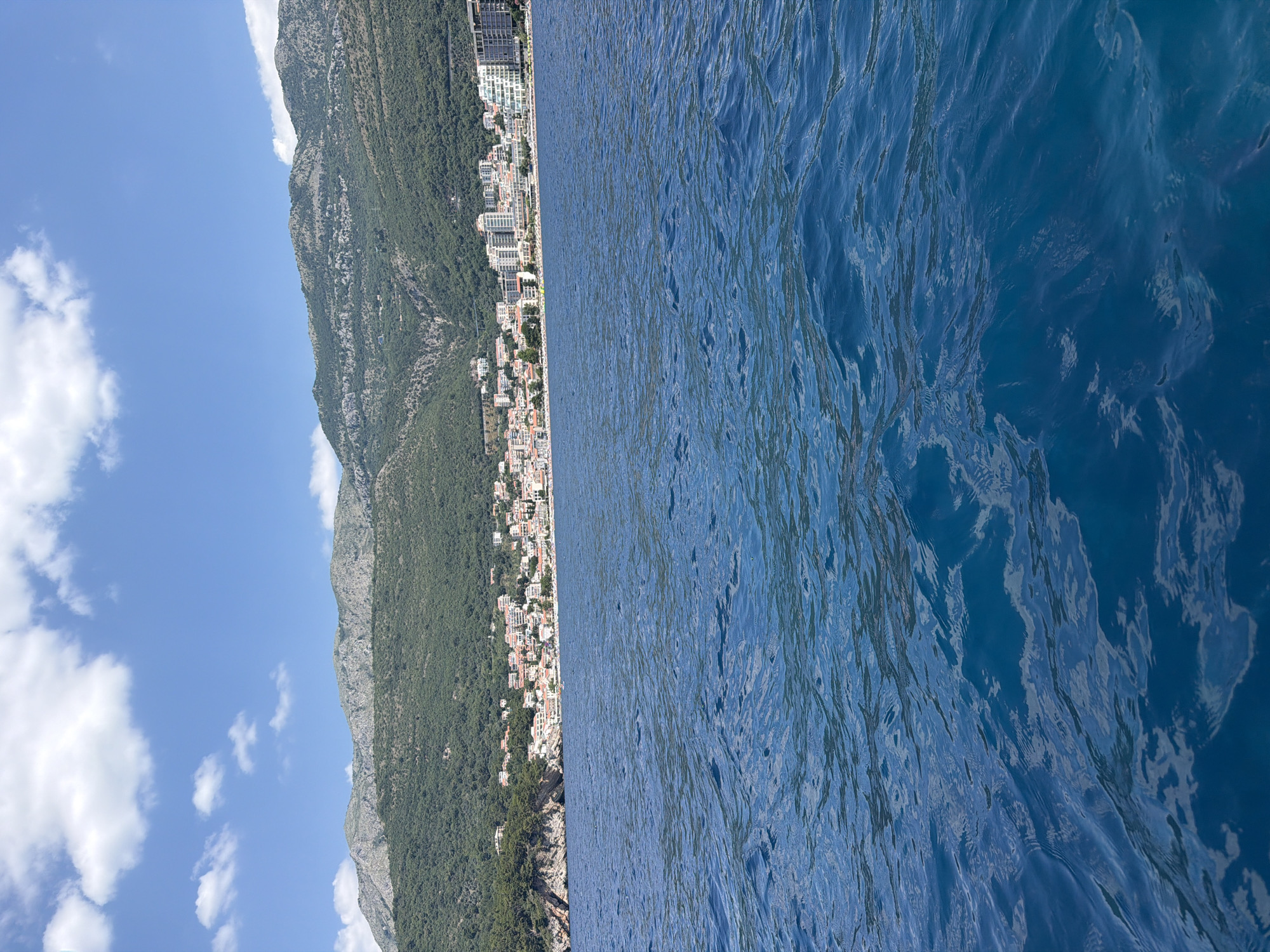 The image captures a scenic view of Petrovac centar, Montenegro, from a body of water. The foreground is dominated by the deep blue, rippling surface of the sea, reflecting the bright sky. In the midground, a coastal town is visible, characterized by a dense cluster of buildings with red-tiled roofs and modern white structures. Beyond the town, lush green hills rise steeply, forming a mountainous backdrop. The sky above is a vibrant blue, dotted with fluffy white clouds, suggesting a clear, sunny day. The perspective suggests the photo was taken from a boat or a similar vantage point on the water. There are no people or specific actions depicted in the scene, focusing purely on the landscape and the coastal settlement.