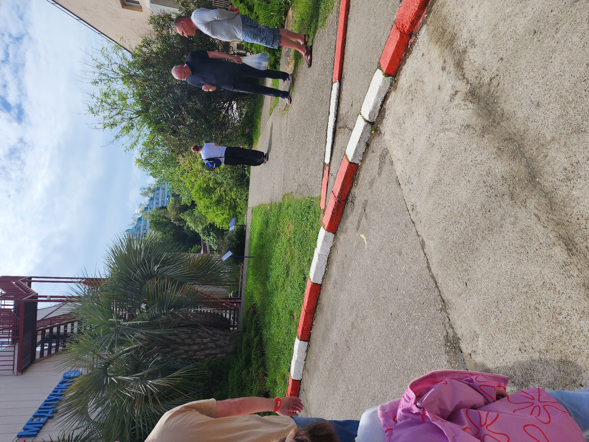The image shows a partially obscured view of a scene in Sochi, Russia. In the foreground, there are parts of people and objects including a torso, jacket, and arm with a red wristband. A red and white curb runs along the edge of a paved pathway. In the background, there are several people walking along the path. To the left, there is a palm tree and a building with blue Cyrillic letters on it. Above the building, there is a red metal staircase. Lush greenery, including trees and bushes, can be seen behind the path. The sky is partly cloudy.