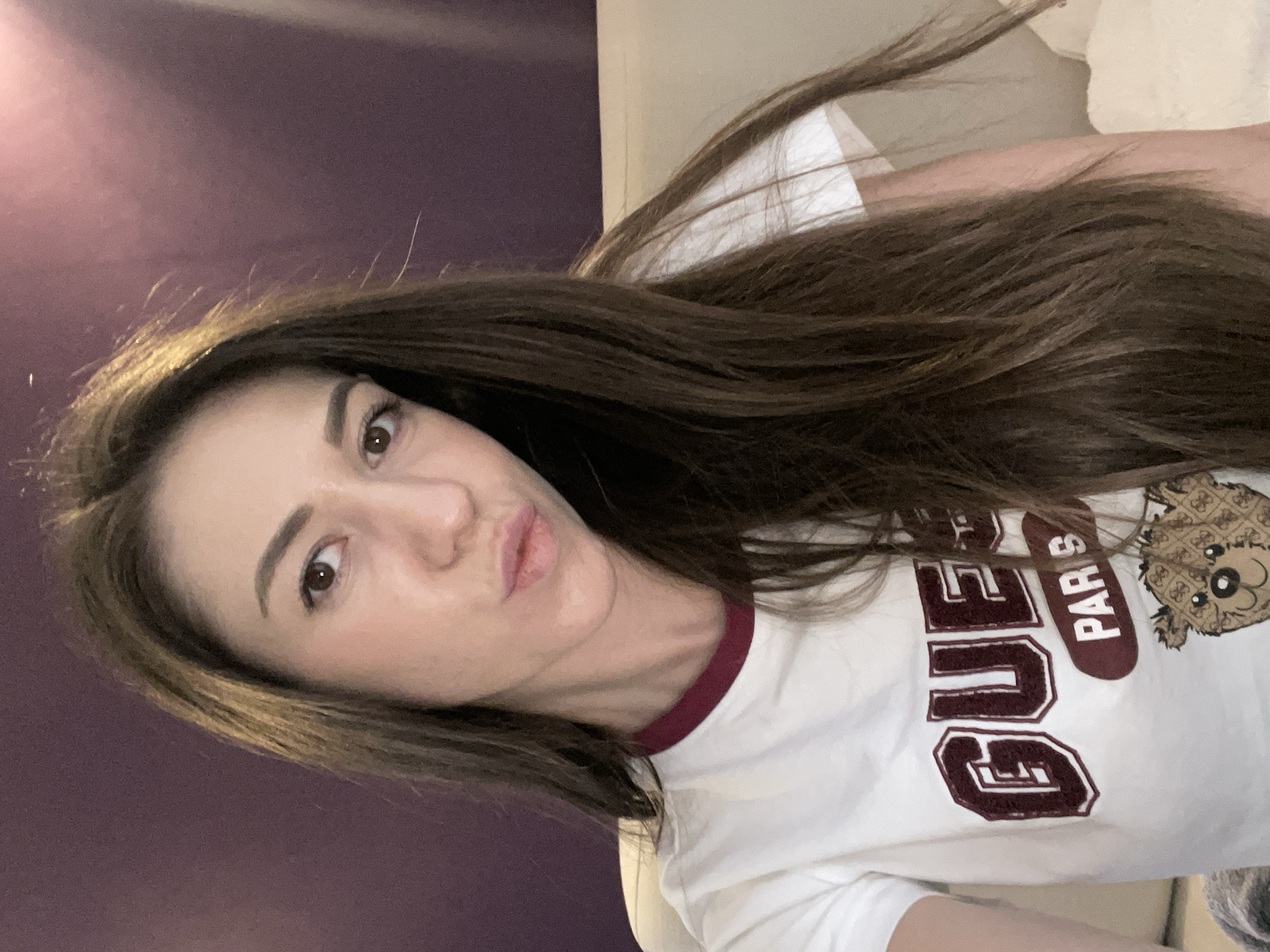A woman takes a close-up selfie, captured at a slight angle with her head tilted to the right. She has long, dark brown hair with lighter brown highlights, which flows across the frame. Her face is turned towards the camera, and she has dark eyes and well-defined eyebrows. Her lips are slightly pursed in a playful expression. She is wearing a white t-shirt with a maroon ringer collar. On the front of the t-shirt, maroon text in a collegiate style reads "QUEEN PARIS," with "PARIS" positioned vertically. Adjacent to the text is a stylized graphic of a cartoon bear's head with a textured pattern, possibly resembling a pineapple or waffle. The background on the left features a dark purple wall with some light reflections, while the right side shows a lighter, off-white or beige surface, suggesting an indoor setting. The image conveys a casual and relaxed atmosphere, likely taken in a room in Sofia, Bulgaria.