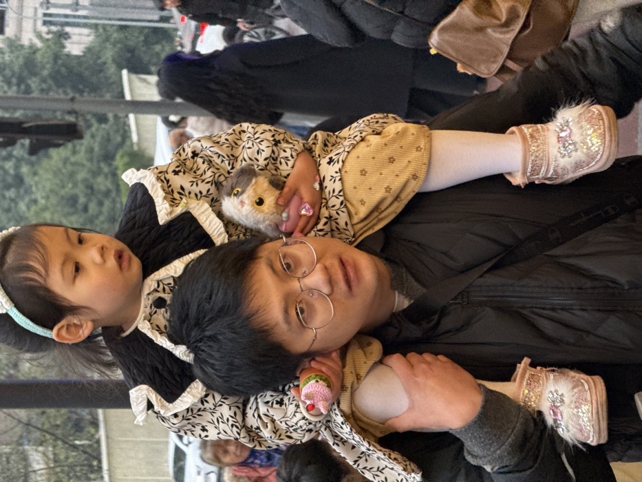 The image captures an East Asian male carrying a young East Asian child on his shoulders in an urban environment in Liangjiang, China. The male subject wears gold-rimmed rectangular glasses, a dark puffer jacket over a grey long-sleeved shirt, and has dark, short hair. He uses both hands to support the child's legs on his shoulders. The child, wearing a light blue knitted headband, a cream-colored dress with a dark leaf pattern, a black quilted collar with white ruffled trim, light brown patterned leggings, white tights, and pink sparkly fur-trimmed boots with a decorative buckle, holds a small grey and white plush toy in her right hand. In her left hand, she holds a small pink knitted ice cream cone toy and wears a prominent pink heart-shaped ring. The male subject looks slightly to his left, while the child looks towards the right side of the frame. The background is an out-of-focus city street scene with blurred pedestrians, vehicles, distant buildings, and a dark traffic light pole, indicating a public outdoor location during daylight.