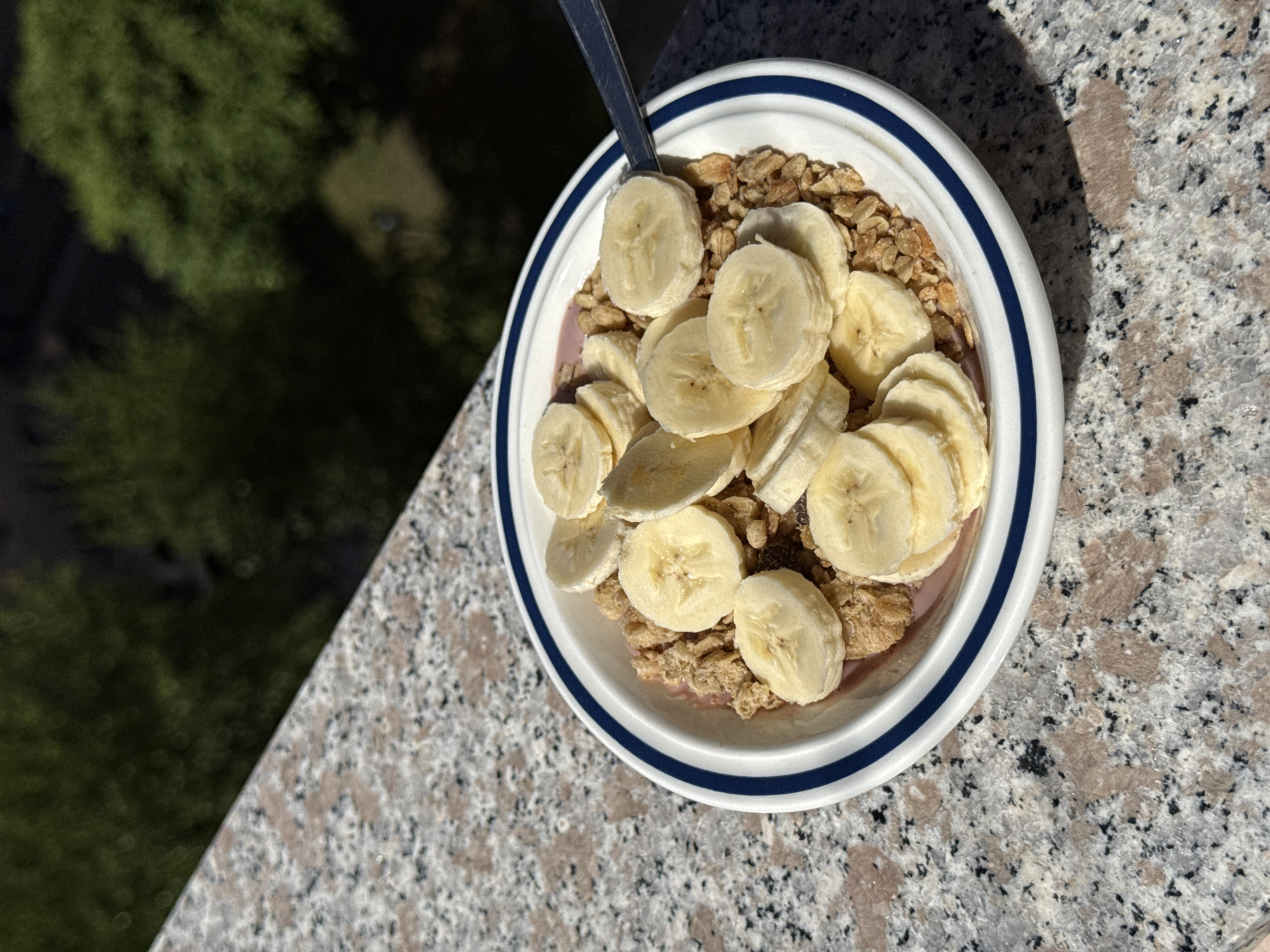 A close-up, high-angle shot captures a bowl of breakfast or a snack placed on a granite-like surface, likely a balcony railing or outdoor table in Milan, Italy. The bowl is white with two thin blue stripes circling its rim. Inside, it contains a generous serving of granola or cereal, topped with numerous slices of fresh banana. A silver spoon is visible, resting in the bowl, suggesting the meal is either about to be eaten or is in progress. Beneath the granola and bananas, a purplish or pinkish layer, possibly yogurt, a smoothie, or fruit puree, is visible.
The surface the bowl rests on is a light gray and white speckled granite with darker black and brown inclusions. Strong, distinct shadows cast by the bowl and the spoon indicate bright, direct sunlight, suggesting it is daytime, likely morning or early afternoon. In the soft-focus background, lush green foliage from trees or bushes is visible, providing a natural contrast to the urban-style stone surface and the prepared food. The scene conveys a peaceful moment of enjoying a meal outdoors under sunny weather. There is no visible text in the image.