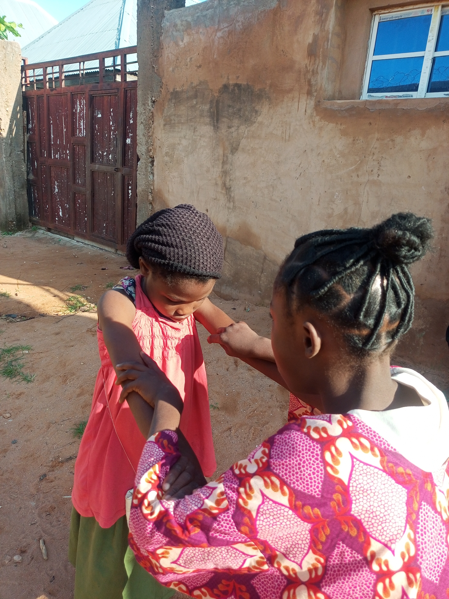 In Tempure, Nigeria, two young girls stand in a dusty, outdoor courtyard. The girl on the left, wearing a dark crocheted hat and a pink dress over a green skirt, looks down at her hands, which are clasped with the other girl's. The girl on the right, facing away from the camera, has her hair styled in braids pulled into a bun at the top of her head. She wears a vibrant pink garment with a white hood, adorned with a bold, patterned design of orange and red flames on a white and pink background. Behind them, a rough, tan-colored wall of a building is visible, featuring a window with blue panes. To the left, a large, dark red metal gate with weathered paint stands partially open, revealing a glimpse of a corrugated metal roof behind it. The ground is covered in dry dirt and scattered with a few small green plants and twigs. The lighting suggests it is daytime, with the sun casting shadows.