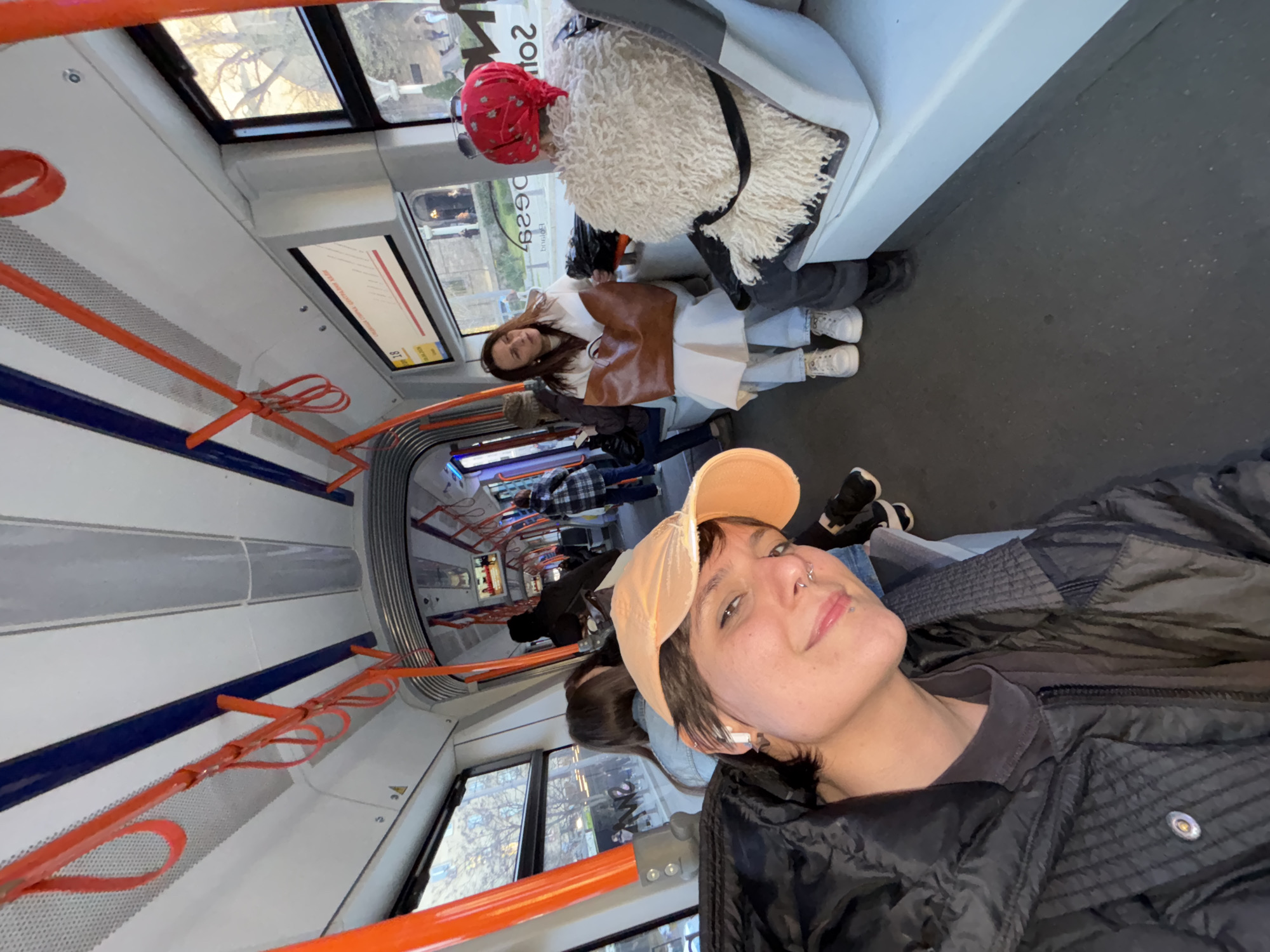 The photo shows the interior of a public transport vehicle, likely a tram or light rail, based on the visible handrails and seating arrangement. The person in the foreground wears a black puffer jacket and a peach-colored baseball cap. The person is looking at the camera and smiling.
In the background, other passengers are visible. A person with a red bandana and a furry white jacket is sitting near the window. Another person in a white coat with a brown vest is seated next to them.
The location is likely in Sofia, Bulgaria.