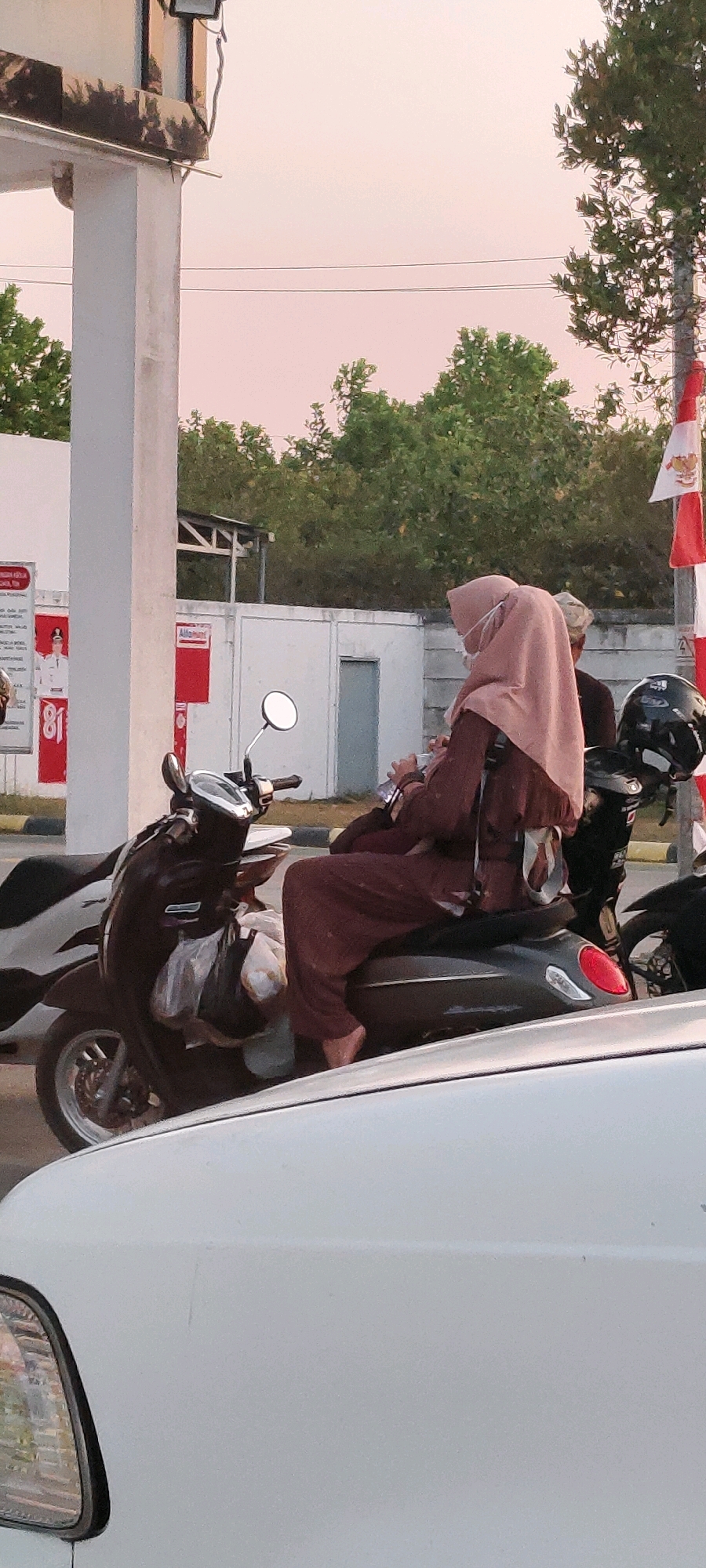 An adult wearing a light pink hijab and a dark brown outfit is seated on a dark grey scooter. They are holding a smartphone in their right hand and appear to be looking at it. Another adult is partially visible behind the scooter rider, wearing a dark shirt. The scooter has a white bag hanging from the handlebars and a black helmet on the back. The scene is set outdoors, with a white pillar and a building in the background. Trees and a pole with red and white Indonesian flags are also visible. The sky is a soft pinkish hue, suggesting it is either dawn or dusk. The image was taken in Bentok Kampung, Indonesia.