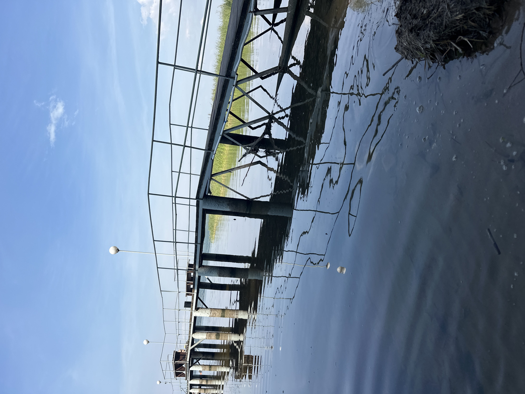 An eye-level, medium shot captures a wooden pier extending into a calm body of water under a bright, partly cloudy sky. The pier, made of dark wood planks with a light-colored metal railing, is supported by numerous concrete pillars. The pillars create elongated, dark reflections on the water's surface, which is disturbed by small ripples and scattered debris. The scene is set in Staromainskii Zagotskot(Nezhil), Russia, with reeds visible along the far bank of the water, suggesting a natural or semi-natural environment. The lighting indicates it is daytime, likely late morning or early afternoon. There are no people or animals present in the image.