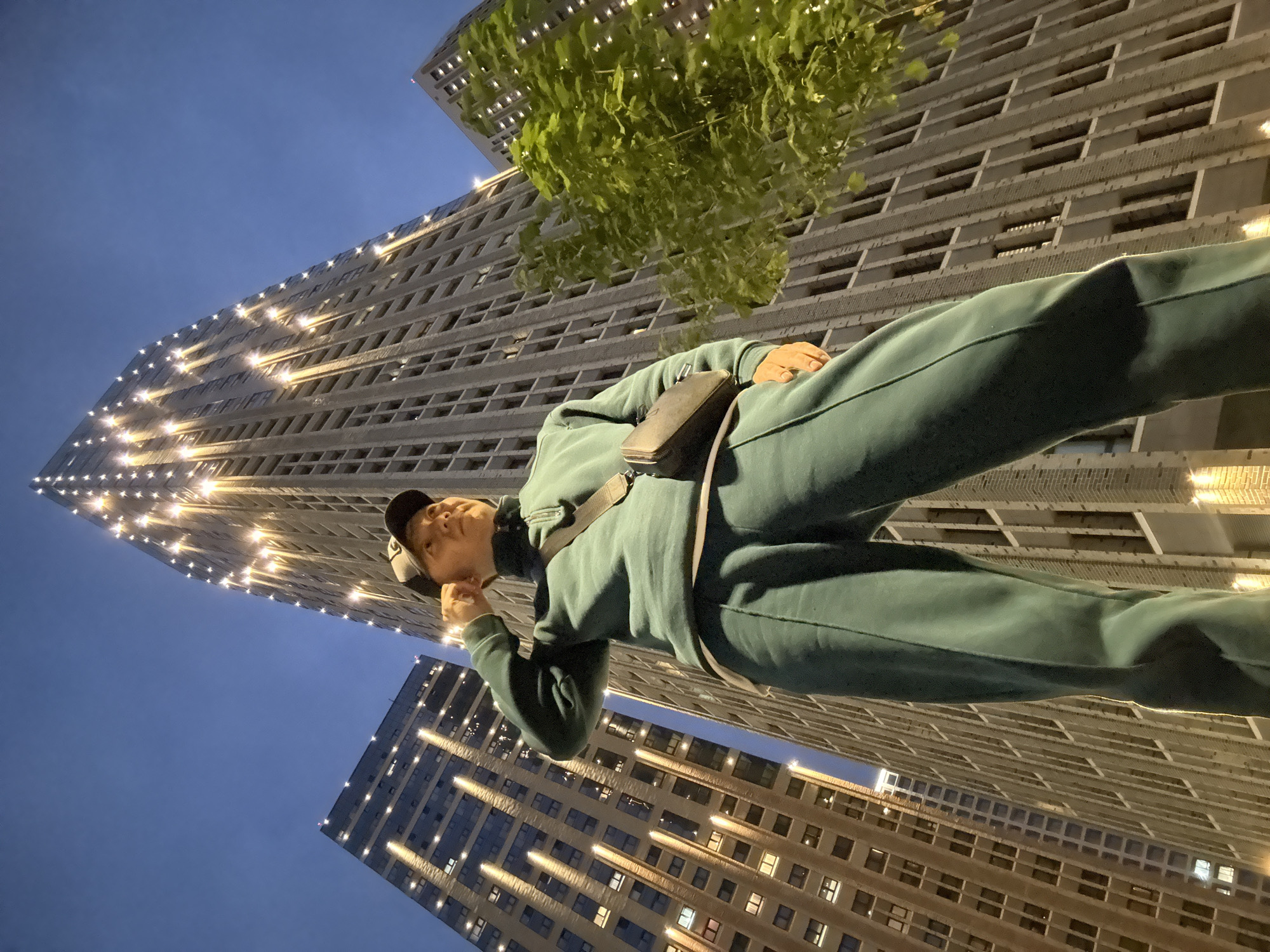 A low-angle shot captures a person in a teal tracksuit standing outdoors at night, facing a row of illuminated modern buildings in Moscow, Russia. The person, wearing a dark baseball cap and a cross-body bag, has their right hand raised to their ear. The buildings, with their many windows and vertical lighting, dominate the background against a deep blue sky. A green leafy tree is visible on the right side of the frame.