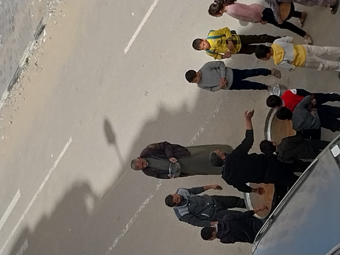 This image, captured in Khan Yunis, Palestinian Territory, depicts an outdoor scene on a paved street, viewed from an elevated perspective with a dark vehicle partially visible in the lower foreground. Approximately twelve individuals are gathered, consisting of at least three adult males and numerous children of varying ages.
At the center of the gathering are two large, dark metal pots or cauldrons, placed directly on the pavement, each containing a substantial quantity of a light brown, semi-solid food. An older adult male with a grey beard, wearing a dark jacket over a light-colored garment, stands holding a silver metal bowl with both hands. To his left, another adult male wears a grey and black jacket. A third adult male, wearing a black hoodie and mostly facing away from the camera, gestures with his right hand towards one of the large pots.
Approximately eight to ten children are visible, standing around the large pots. They are dressed in various casual attire, including a boy in a yellow jacket with a backpack, another in a red shirt, and others in darker or lighter clothing. Some children are near or appear to be holding smaller metal bowls. The individuals are clustered around the food pots, indicating a communal food distribution or serving event. White road markings are visible on the pavement, and a dirt shoulder with some debris lines the far right.