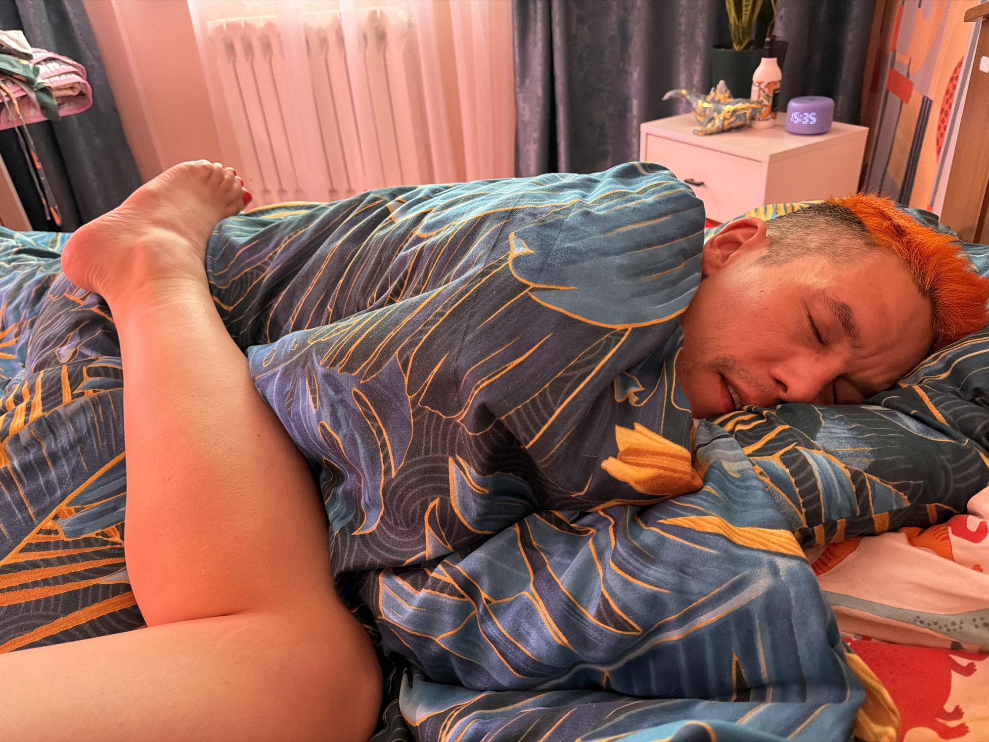 A person with orange hair is asleep in a bed covered with a blue and yellow patterned duvet. The person's left leg, with red nail polish on the toes, is sticking out from under the covers. The location is possibly a residence in Chaikovsky, Russia. A white nightstand is visible in the background with a decorative dinosaur figurine, a lotion bottle, and a digital clock displaying "15:35". A radiator is situated near a window with sheer curtains. A partial view of a clothes rack with hanging garments is present. The scene appears to be a domestic setting.