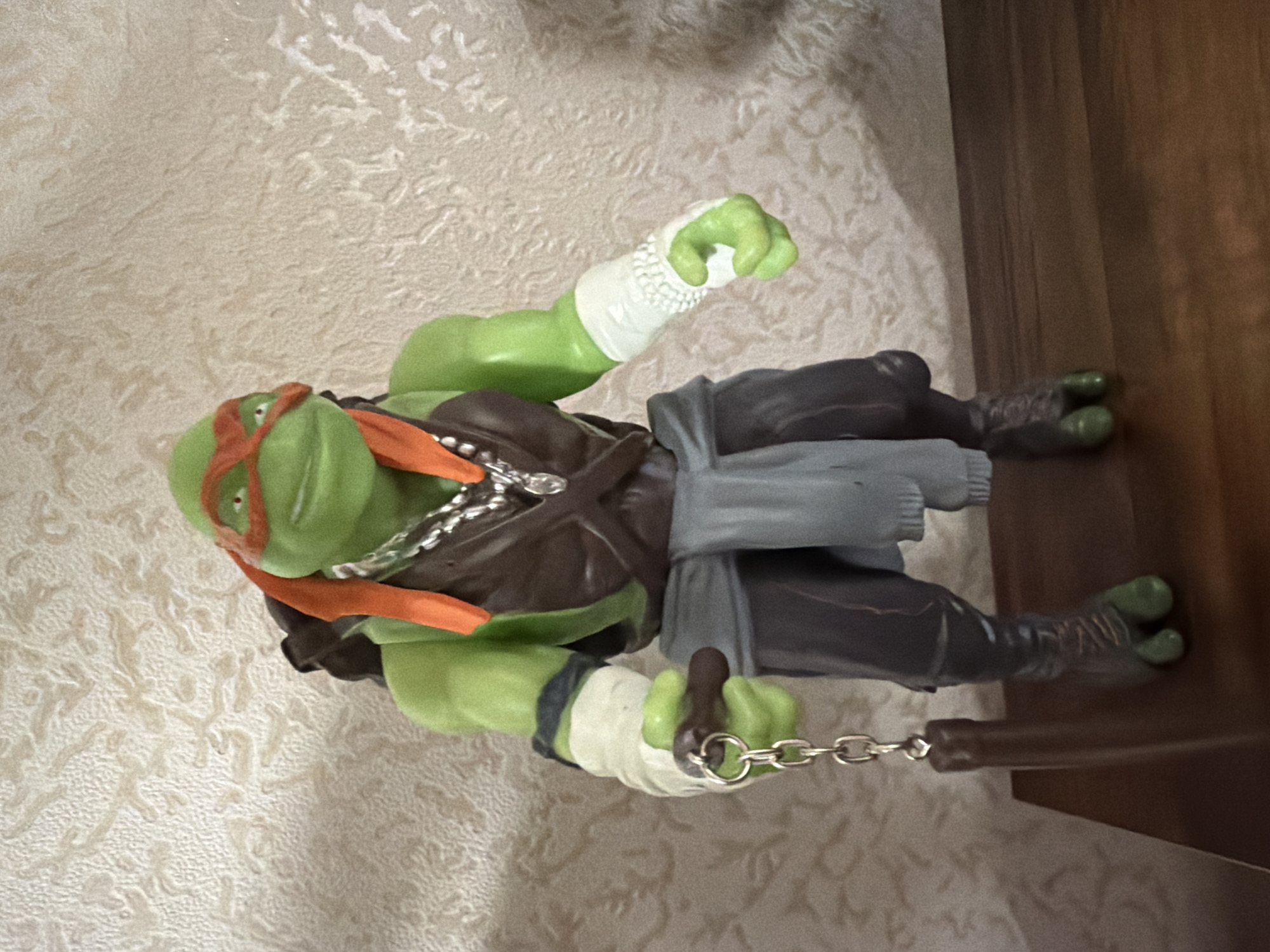 A Teenage Mutant Ninja Turtle action figure, specifically Michelangelo, is standing on a wooden surface against a textured, light-colored wall. The figure is green with an orange bandana and mask, wearing a brown vest, gray sash, and dark pants. It has white bandages on its arms and is holding a nunchaku in its left hand. The figure appears to be in a display or on a shelf. The image does not provide any information about the city of Yablonovskiy, Russia.