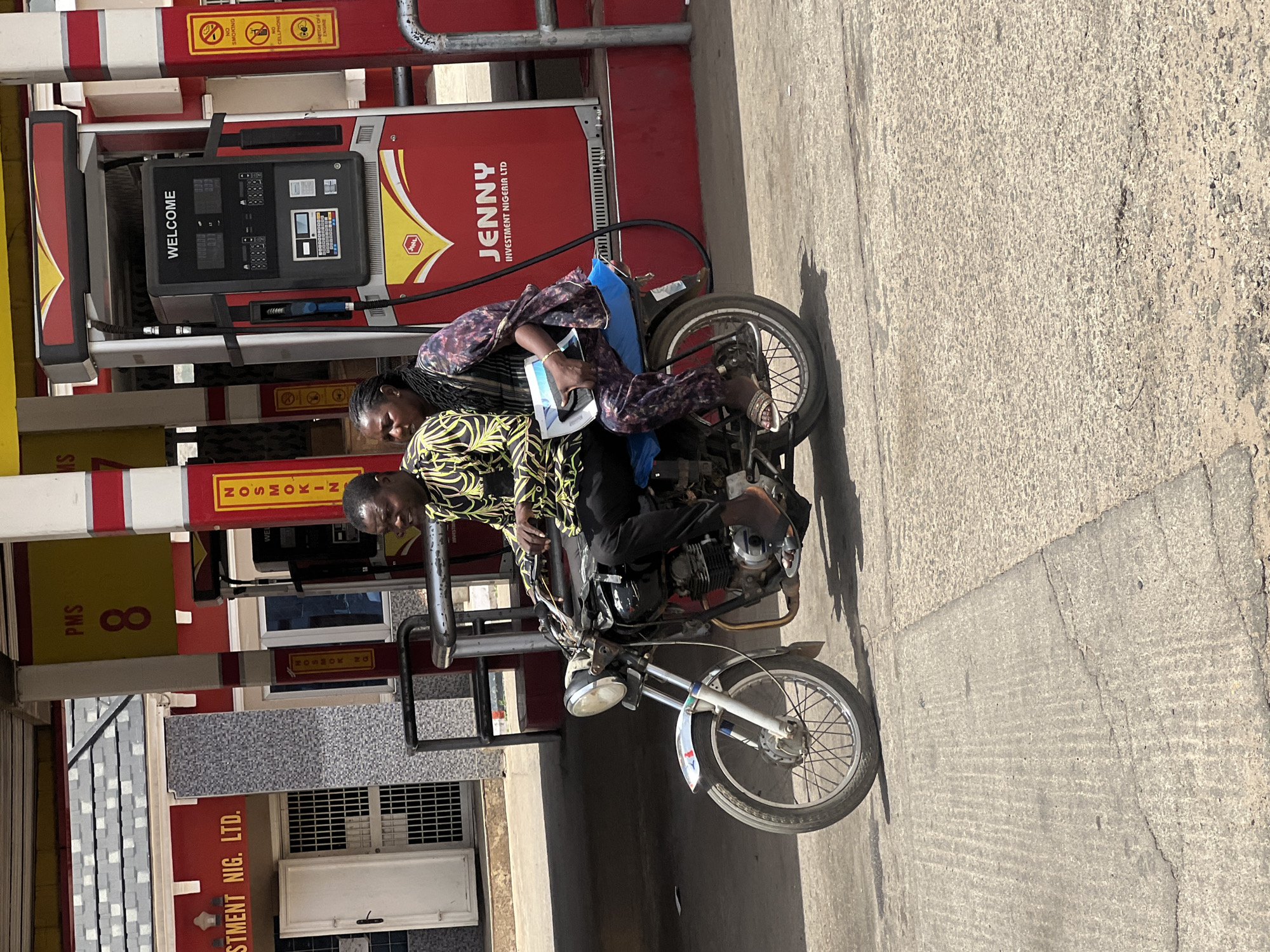 This image, taken in Jos, Nigeria, captures two individuals on a motorcycle at a gas station during bright daylight. A man, wearing a yellow, green, and black patterned shirt, is seated as the driver of the dark-colored motorcycle. A woman, dressed in a purple and white patterned outfit, is seated behind him, holding a white object on her lap. Both individuals appear to be looking forward, past the gas pump.
The setting is a gas station, with a prominent red and black fuel pump visible behind the motorcycle. The pump's digital display shows the word "WELCOME." Directly above the pump, various safety notices are visible, including "NO SMOKING," "NO CELLPHONE," and "SWITCH OFF ENGINE." The gas station branding, "JENNY INVESTMENT NIGERIA LTD," is clearly displayed on the side of the pump. Other partial text visible includes "PMS 8," "STMENT NIG. LTD.," and what appears to be "HOSMOKIN" (likely part of a "NO SMOKING" sign).
The ground is paved, possibly concrete or asphalt, with strong shadows suggesting a sunny day. The scene depicts a routine activity at a fuel station.