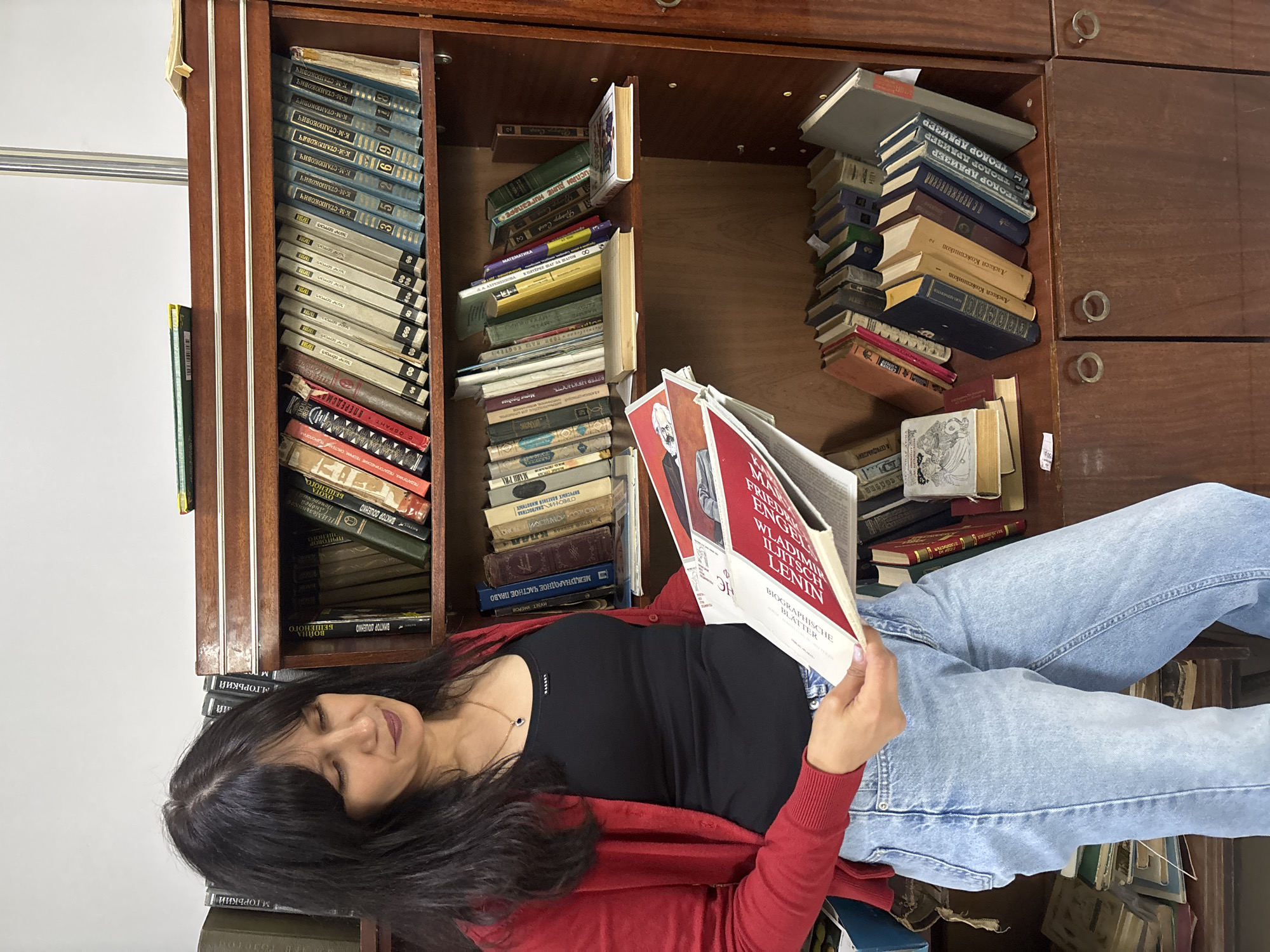 A woman with dark hair, wearing a black top, a red cardigan, and blue jeans, is seen engrossed in reading a book. She holds the book open, her gaze directed at the pages, with a calm and focused expression.
The scene is set indoors, against a large, dark wooden bookshelf or cabinet overflowing with books. The books are arranged densely, some neatly stacked, others leaning or piled haphazardly, indicating a personal and well-used collection. Many books feature Cyrillic script on their spines, suggesting a significant portion of the collection is in Russian.
The book the woman is reading has a red cover and features prominent text in German: "VLADIMIR ILJITSCH LENIN", "FRIEDRICH ENGELS", and "BIOGRAPHISCHE BLÄTTER" (Biographical Pages). Other visible titles and authors on the shelves, identified from the OCR, include multiple volumes by "К-М-СТАНЮКОВИЧ" (K-M-Stanyukovich), "ТЕОДОР ДРАЙЗЕР" (Theodore Dreiser), "М. ГОРЬКИЙ" (M. Gorky), "Алексей Кожевников", "А-СЕРАФИМОВИЧ" (A-Serafimovich), and "ВИКТОР ДОШЕНКО" (Viktor Doshenko). Further titles include "МЕЖДУНАРОДНОЕ ЧАСТНОЕ ПРАВО" (International Private Law), "СПРАВОЧНИК ДИАГНОСТИКА ВИРУСНЫХ БОЛЕЗНЕЙ ЖИВОТНЫХ" (Handbook for Diagnosis of Viral Diseases of Animals), "ВЕТЕР НЕЖНОСТЬ" (Wind Tenderness), and "ИСЛАМ ДИНЕ нигезпе" (Islam Religion Foundation, likely Tatar). The environment suggests a domestic setting in Chaikovsky, Russia.