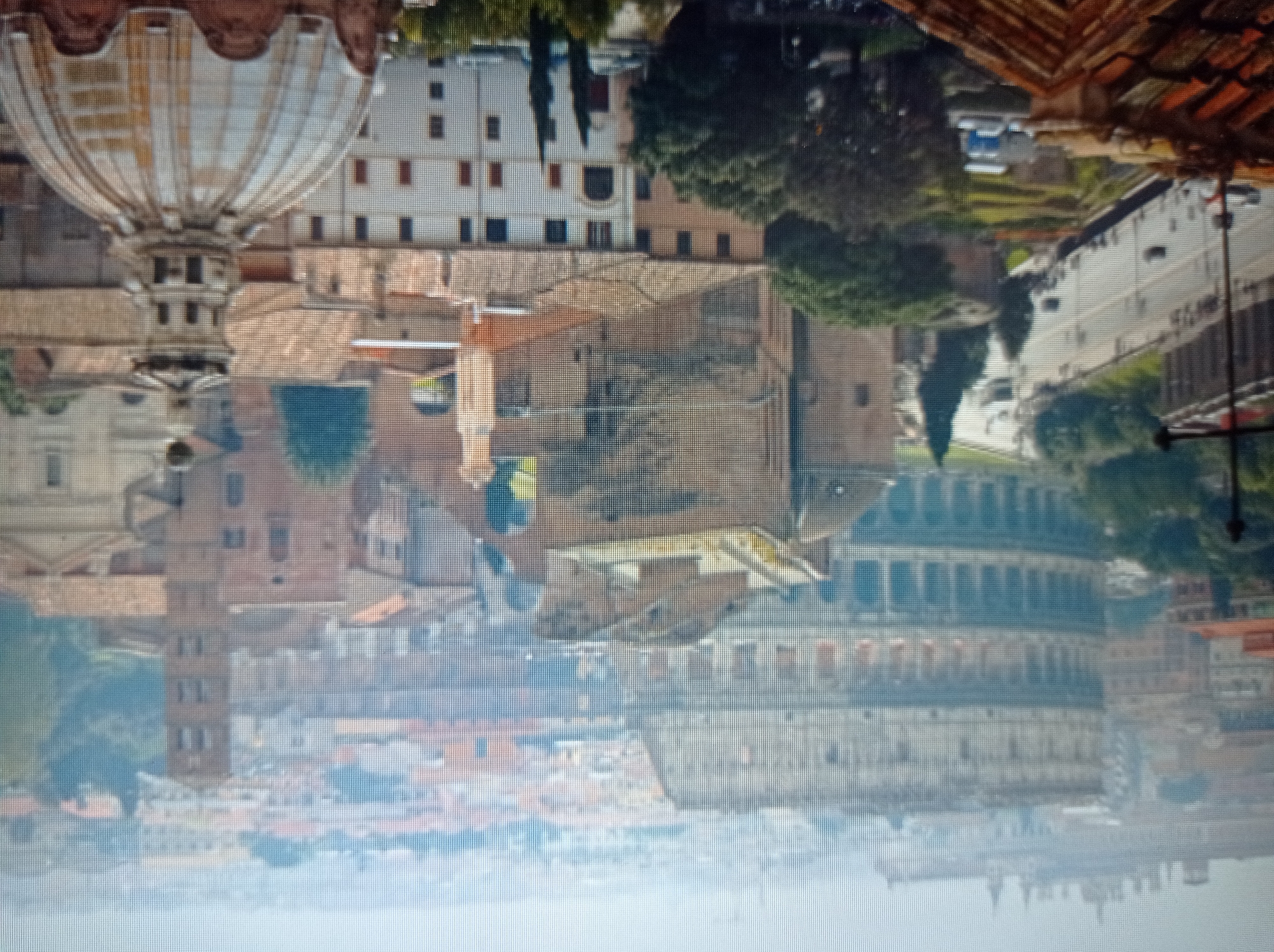 This is a pixelated, aerial photograph of Rome, Italy, featuring the iconic Colosseum prominently in the background. The image captures a dense urban landscape with numerous buildings, rooftops, and some greenery. In the foreground, a domed structure and the upper portions of other buildings are visible. A street with moving vehicles and pedestrians can be seen to the left, bordering a landscaped area. The background shows more of the cityscape, with a tall tower structure to the right of the Colosseum. The atmosphere appears slightly hazy, suggesting a distant view. The image does not depict Mukachevo, Ukraine.
