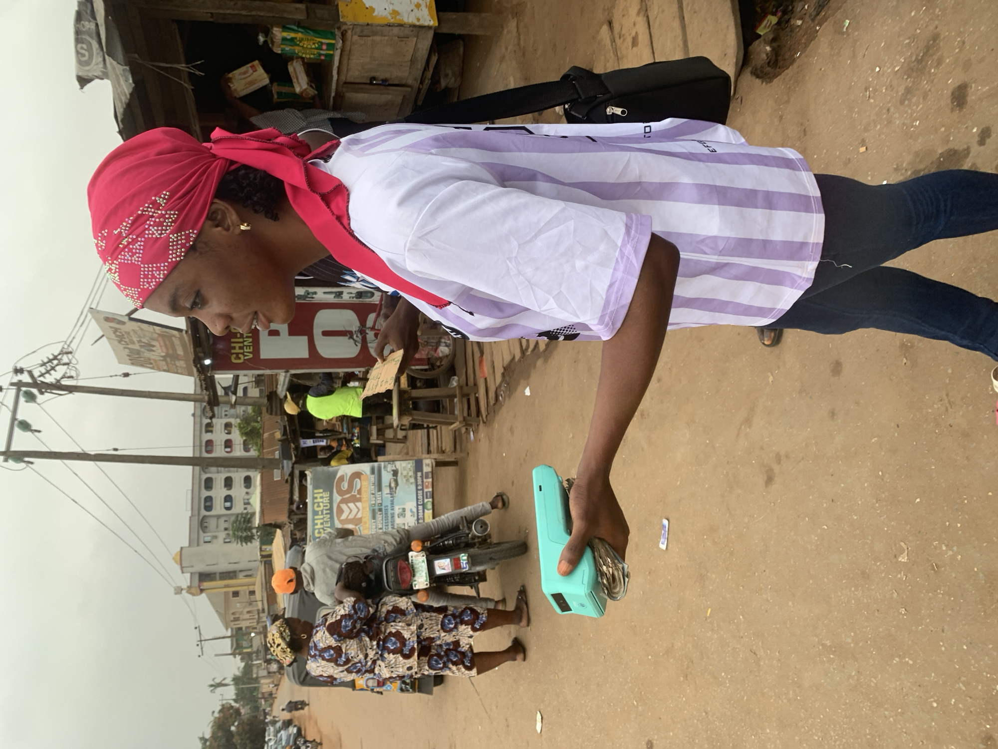 A young Nigerian woman, wearing a pink headscarf and a white and purple striped t-shirt, stands on a dirt road in Shagamu, Nigeria, holding a teal payment terminal and a wad of cash. She is in the foreground, facing right in profile, with her right arm extended holding the payment device. In the background, several people are gathered around a motorbike and a market stall. The environment appears to be a bustling street scene with buildings, utility poles, and other market structures.