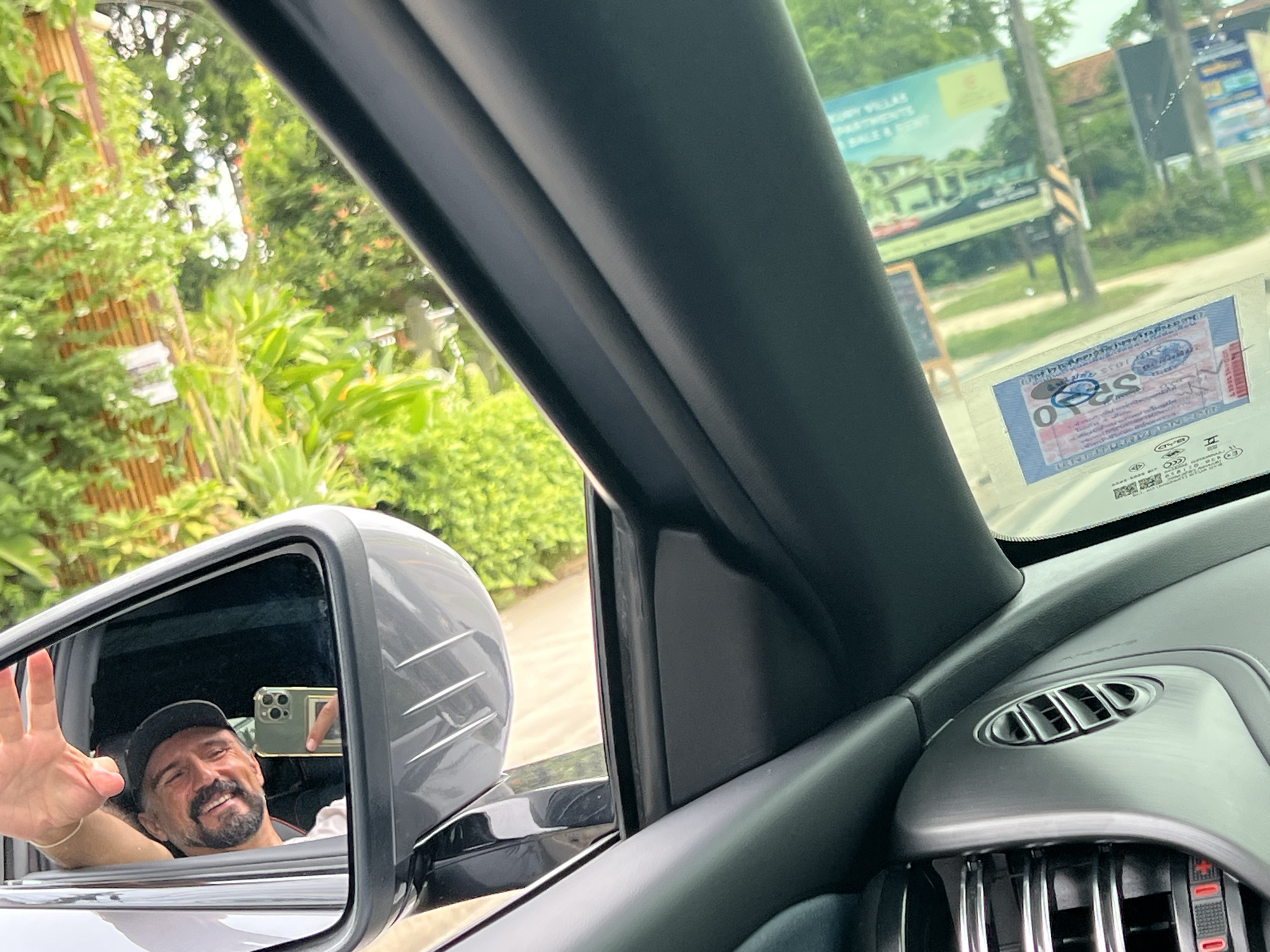 A middle-aged man with a beard and a cap is reflected in a car's side mirror, waving with his left hand. He is holding a smartphone in his right hand, which is angled to capture the reflection. The man appears to be inside a vehicle, possibly driving or as a passenger. The background visible in the mirror shows lush green vegetation, suggesting an outdoor setting. Through the car's windshield, further in the distance, a billboard with text and images of houses is visible, along with streetlights and more greenery. The car's interior is also partially visible, with a black dashboard and an air vent. Based on the context of the billboard, the location is identified as Baan Lamai, Thailand. The image is taken from inside the car, looking out.