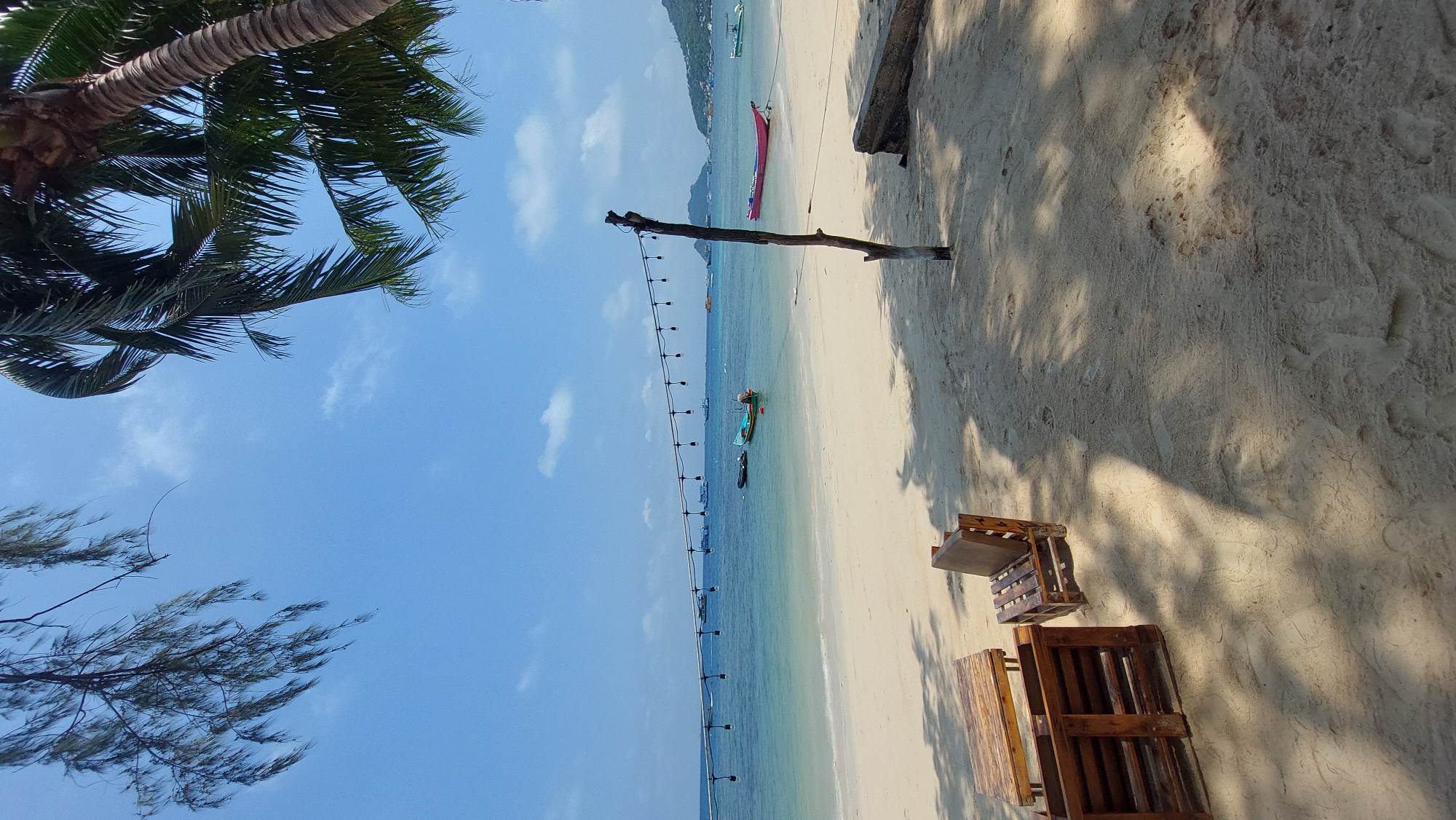 The image depicts a beach scene in Tambon Ko Tao, Thailand. The perspective is a slightly rotated, ground-level view. In the foreground, there are wooden pallet chairs on the sand. The sand itself is light in color and shows various shadows, likely from trees or objects off-frame. The ocean stretches out from the beach, appearing turquoise near the shore and gradually transitioning to deeper blue. Several boats are visible on the water. A string of lights is suspended in the air above the beach. A wooden post is planted in the sand, possibly used to support the string of lights. Trees are present on the left side of the frame, including a palm tree. The sky is a clear blue with scattered white clouds. In the distance, land is visible.