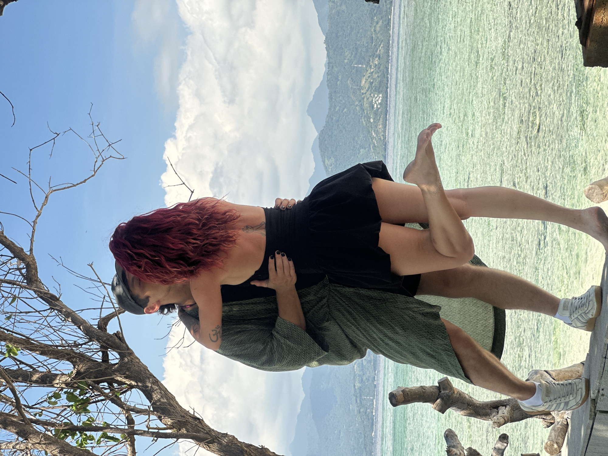 A man and a woman are embracing on an elevated wooden platform near the water in an unknown location in Indonesia. The woman has red hair, a black dress, and visible tattoos on her back and arm. The man is wearing a hat, a patterned green robe, and white sneakers. The background consists of a body of water with mountains in the distance and a partially cloudy sky. Branches of a tree are visible in the lower-left corner of the image.